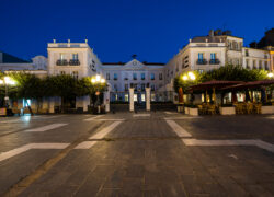 en::U-shaped building seen at night from a square lit by street lamps, lined with trees on either side of the gate and entrance porch. fr::Bâtiment en U vu de nuit depuis une place éclairée par des lampadaires, bordée d’arbres de part et d’autre de la grille et du porche d’entrée.