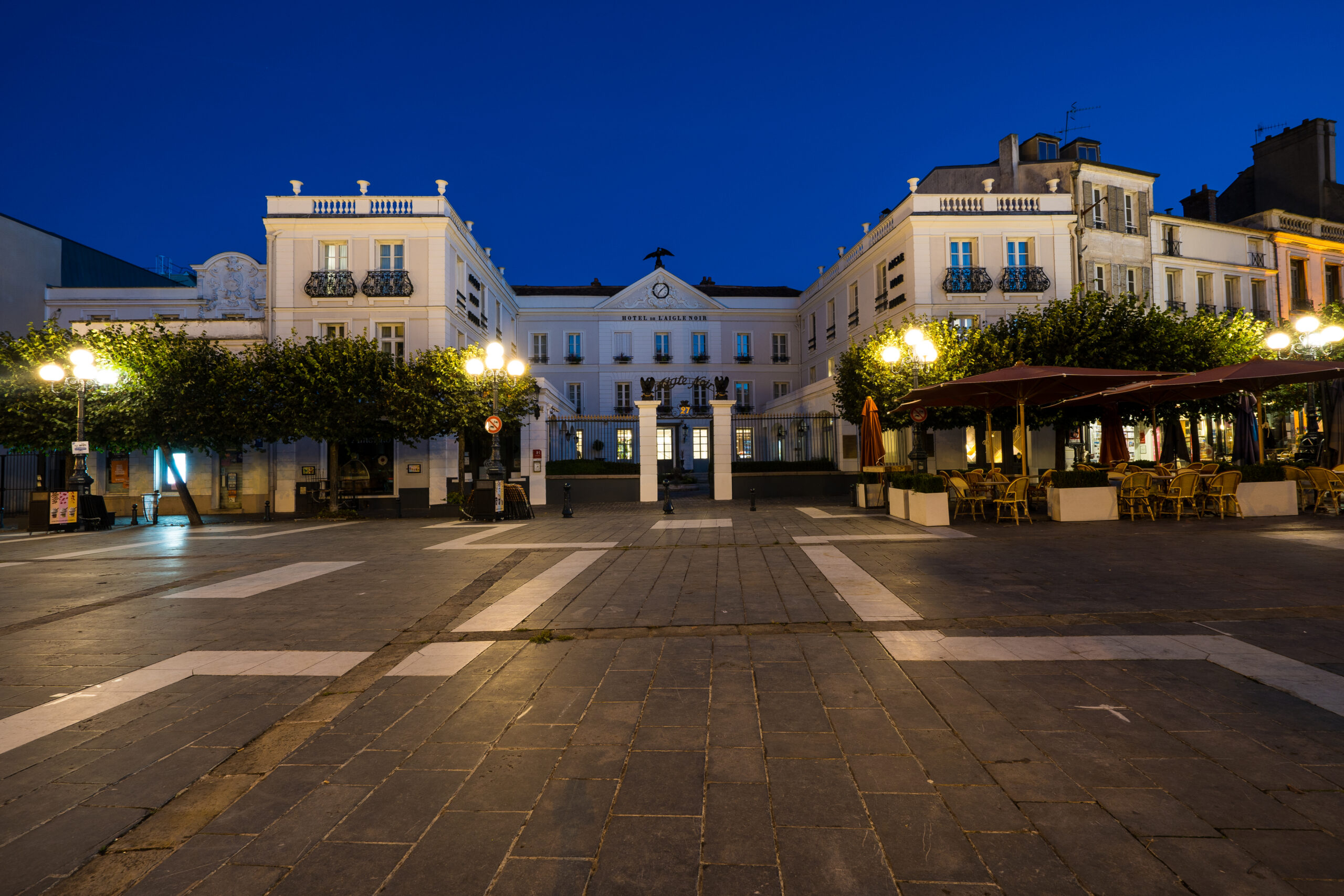 en::U-shaped building seen at night from a square lit by street lamps, lined with trees on either side of the gate and entrance porch. fr::Bâtiment en U vu de nuit depuis une place éclairée par des lampadaires, bordée d’arbres de part et d’autre de la grille et du porche d’entrée.