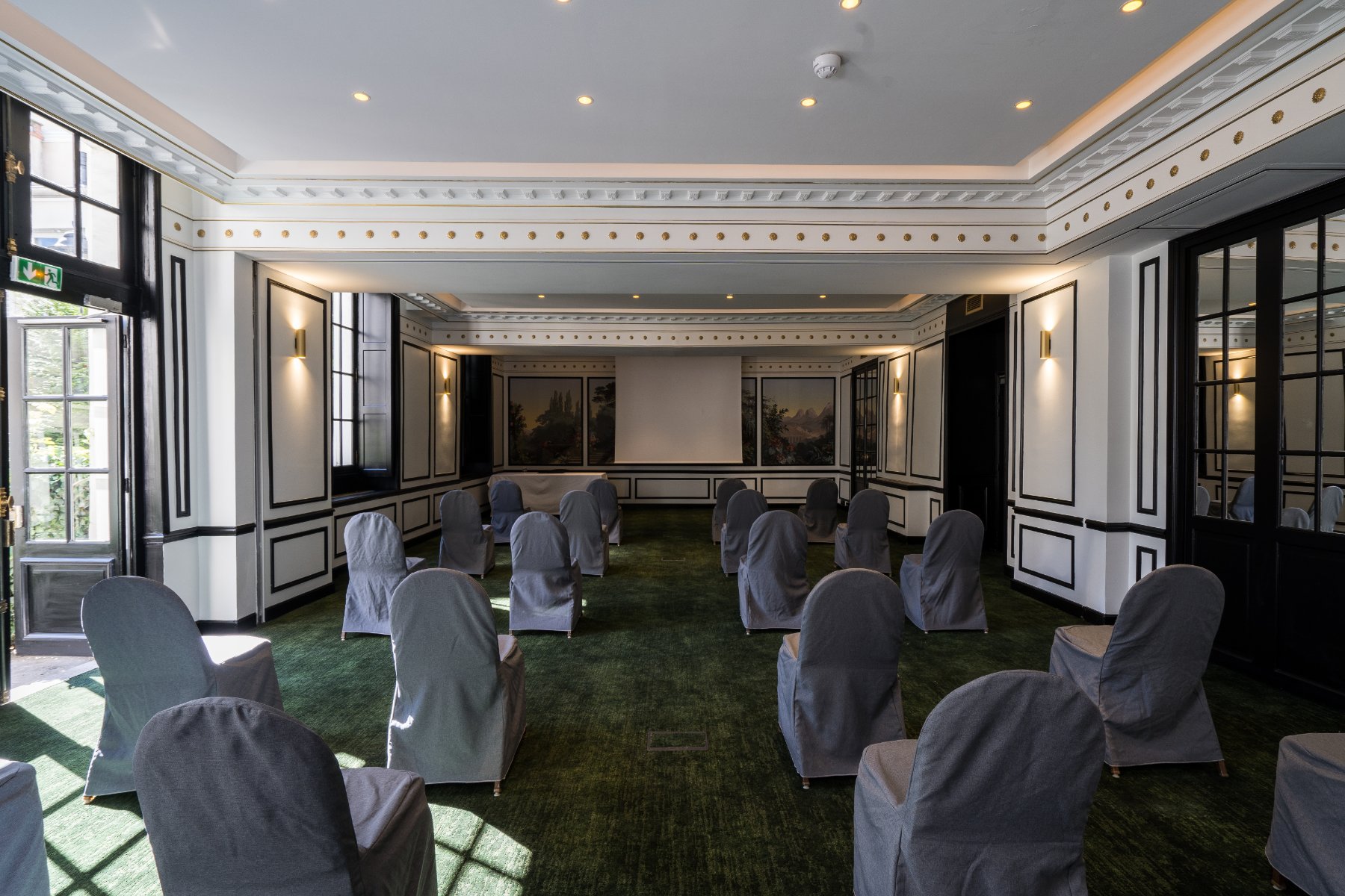 en::Rows of grey fabric armchairs facing a screen, with doors open onto a green inner courtyard. fr::Rangées de fauteuils en tissu gris face à un écran, avec portes ouvertes sur une cour intérieure végétalisée.