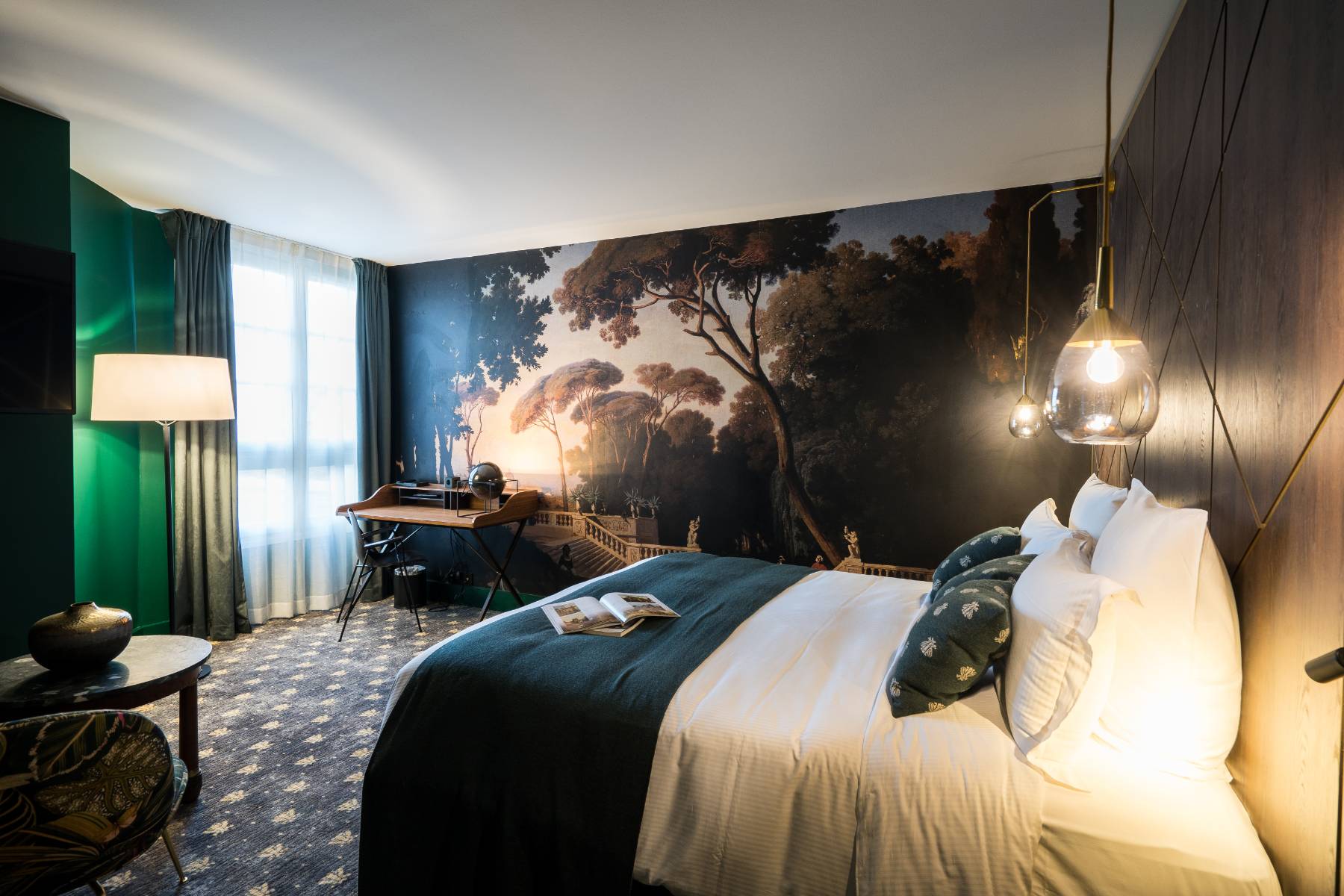 en::Room with an open book on the bed, a tapestry depicting an old staircase surrounded by a large garden, and a secretary desk near the window. fr::Chambre avec livre ouvert sur le lit, tapisserie représentant un escalier ancien entouré d’un vaste jardin, et bureau secrétaire près de la fenêtre.