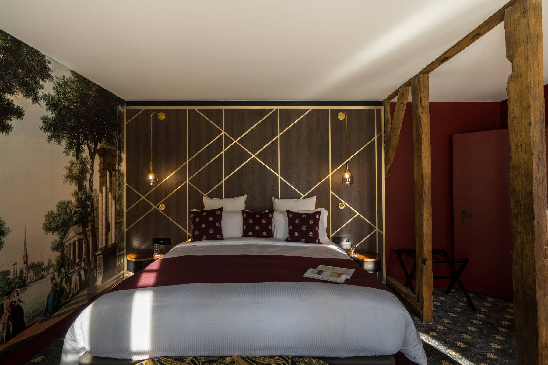 en::Room with exposed beams separating the sleeping and lounge areas, a wooden headboard extended by a tapestry depicting city scenes from the royal era. fr::Chambre avec poutres apparentes séparant l’espace nuit et le salon, tête de lit en bois prolongée par une tapisserie représentant des scènes de ville à l’époque royale.