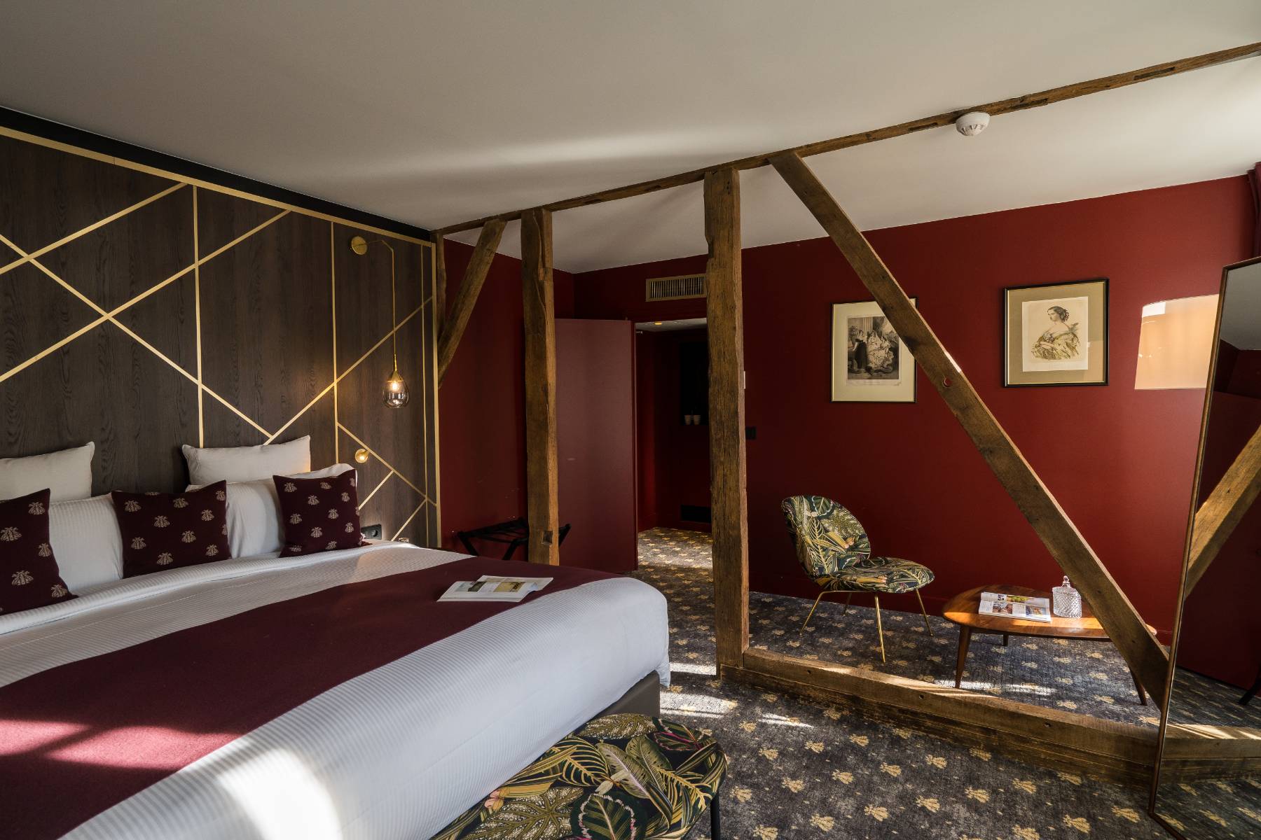 en::Room with exposed beams separating the sleeping and lounge areas, in burgundy, wood and gold tones, featuring a large freestanding mirror. fr::Chambre avec poutres apparentes séparant l’espace nuit et le salon, dans des tonalités bordeaux, bois et or, avec un grand miroir sur pied.