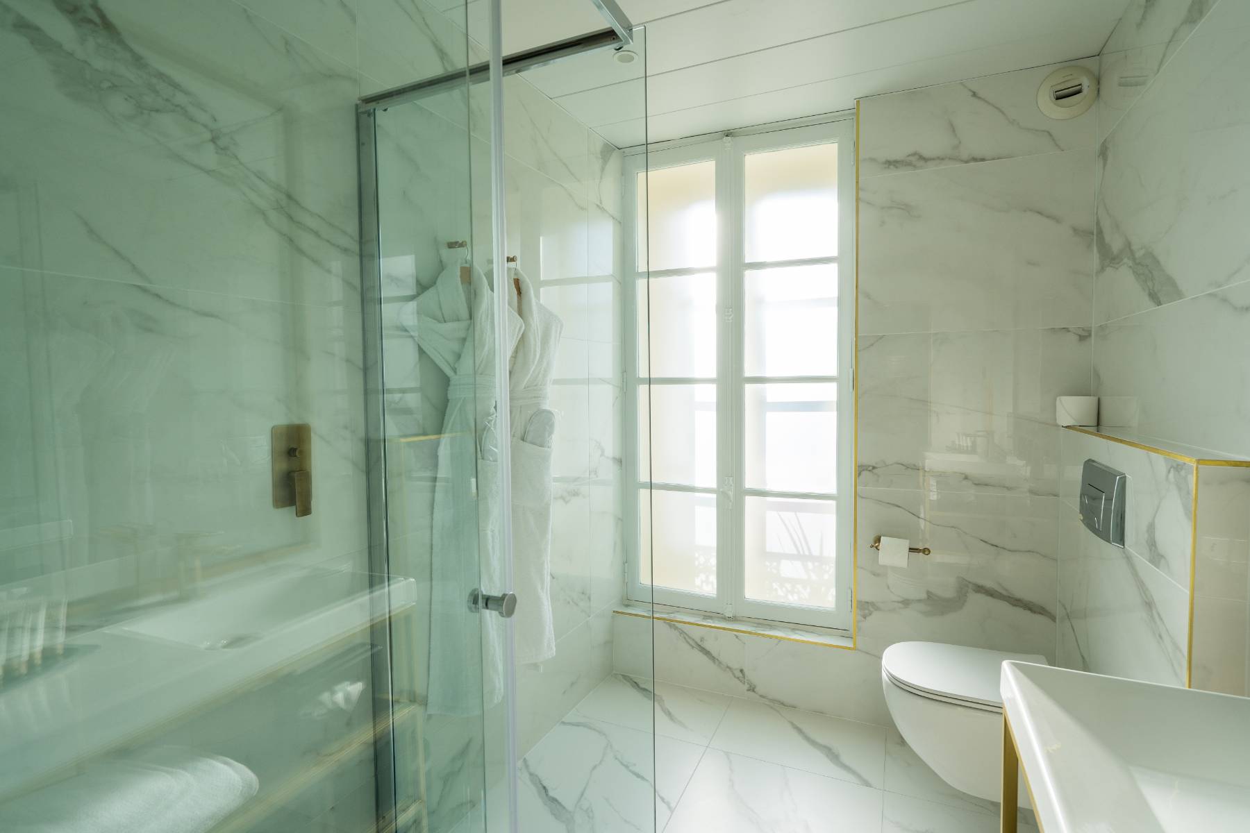 en::Bathroom in white and grey marble tones, with gold finishes and hanging bathrobes. fr::Salle de bain aux tons marbrés blanc et gris, avec finitions dorées et peignoirs suspendus.