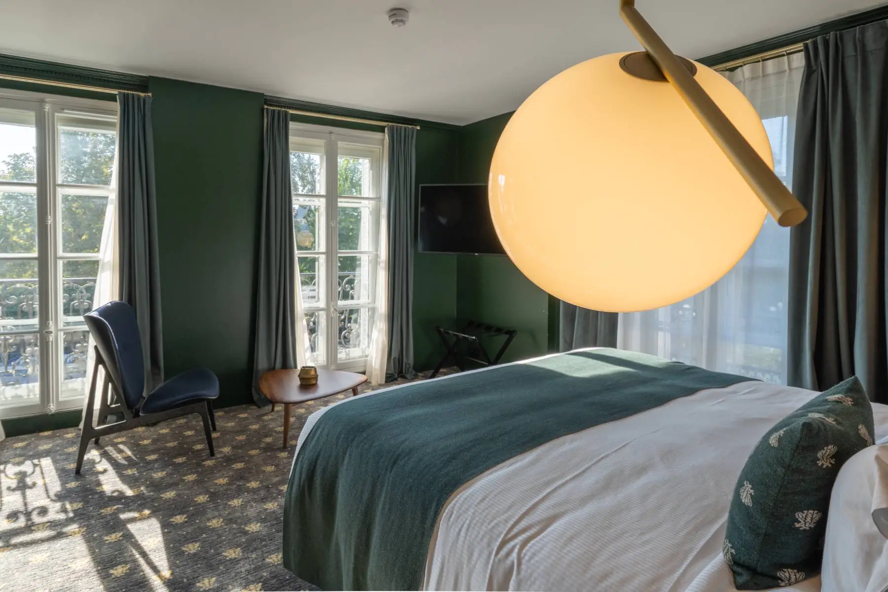 en::Double bed with green bedspread and cushions featuring the hotel’s signature pattern, three large French windows opening onto greenery, a wooden coffee table, and a blue armchair. fr::Lit double avec dessus de lit et coussins verts aux motifs signature de l’hôtel, trois grandes portes-fenêtres ouvertes sur la végétation, table basse en bois et fauteuil bleu.