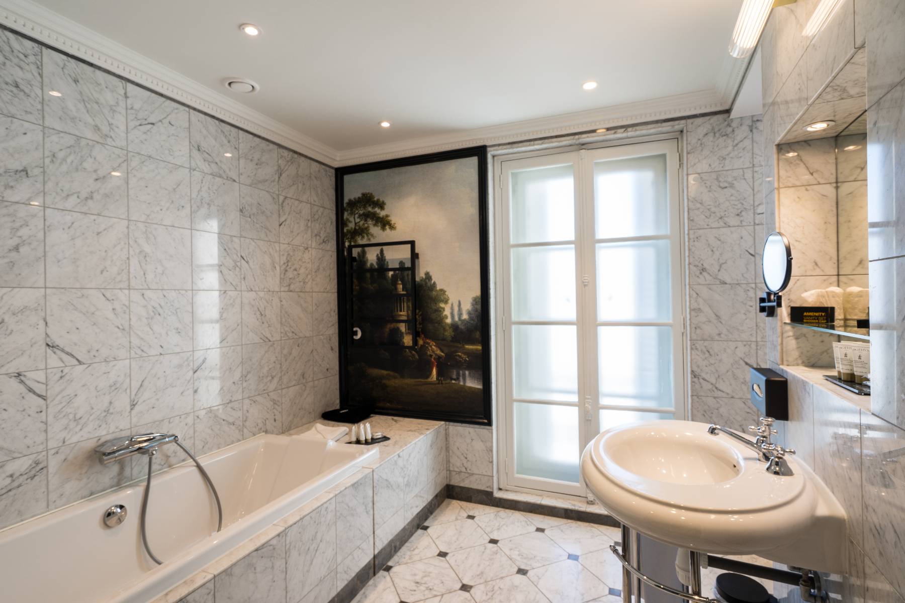 en::Marble bathroom with a landscape wall tapestry, French windows, and a round white sink. fr::Salle de bain en marbre avec tapisserie murale paysagère, portes-fenêtres, et lavabo rond blanc.