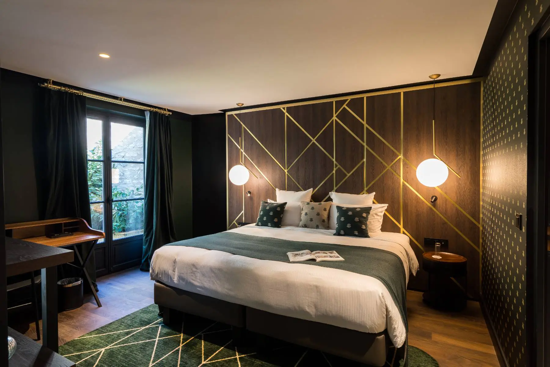 en::Room with a French window opening onto a green outdoor space, bed framed by a wooden wall and a wallpapered wall, carpeted floor, and a secretary desk. fr::Chambre avec porte-fenêtre ouvrant sur un espace végétalisé, lit encadré par un mur en bois et un mur tapissé, tapis au sol et bureau secrétaire.