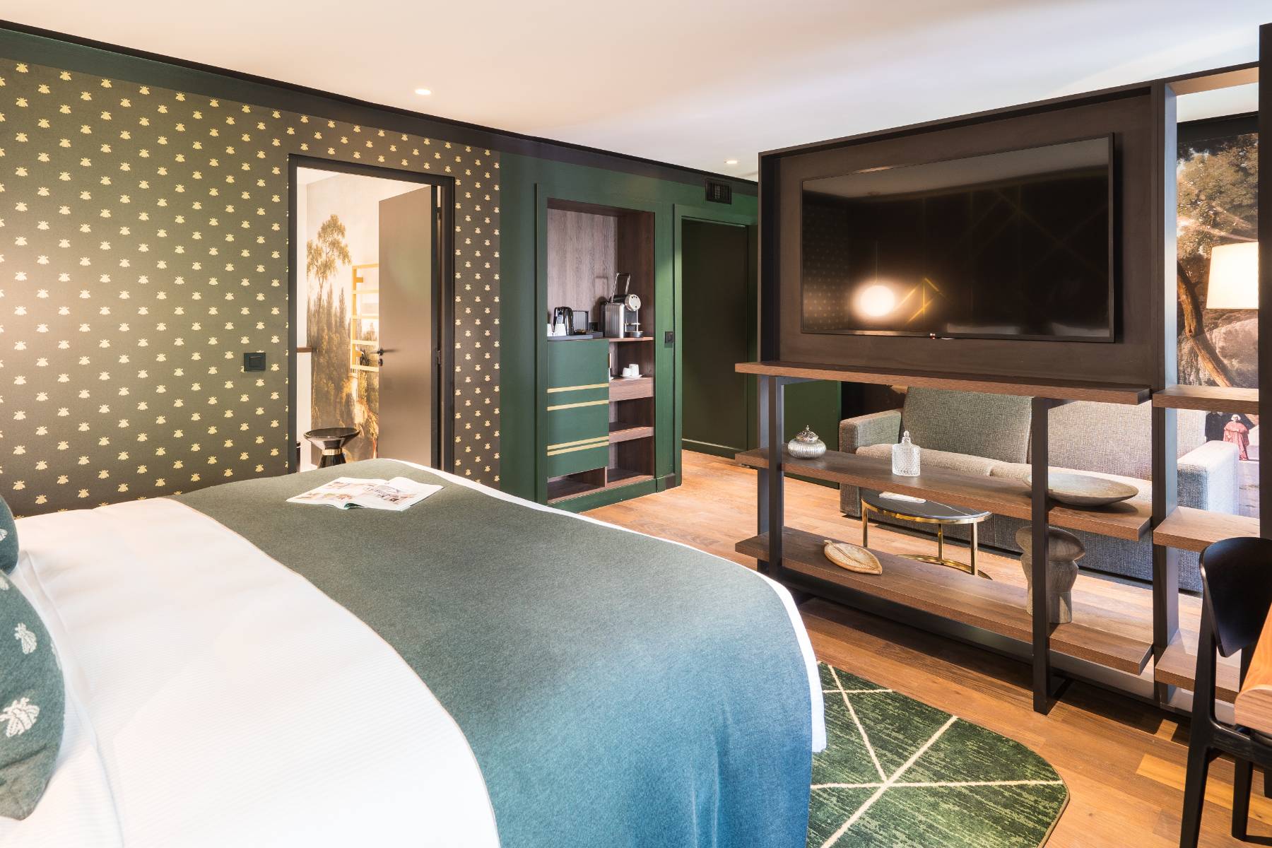 en::Suite viewed from the bed, showing a marble bathroom with wall tapestry, welcome drinks area with coffee machine, and a lounge corner with sofa and shelves displaying vintage objects. fr::Suite vue depuis le lit, avec salle de bain en marbre et tapisserie murale visible, espace boisson avec machine à café, coin salon avec canapé et étagères décorées d’objets anciens.