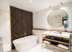 en::Bathroom combining wood, marble and brass, with outdoor greenery reflected in the main mirror and magnifying mirror. fr::Salle de bain mêlant bois, marbre et laiton, avec vue sur la végétation extérieure reflétée dans le miroir central et le miroir grossissant.