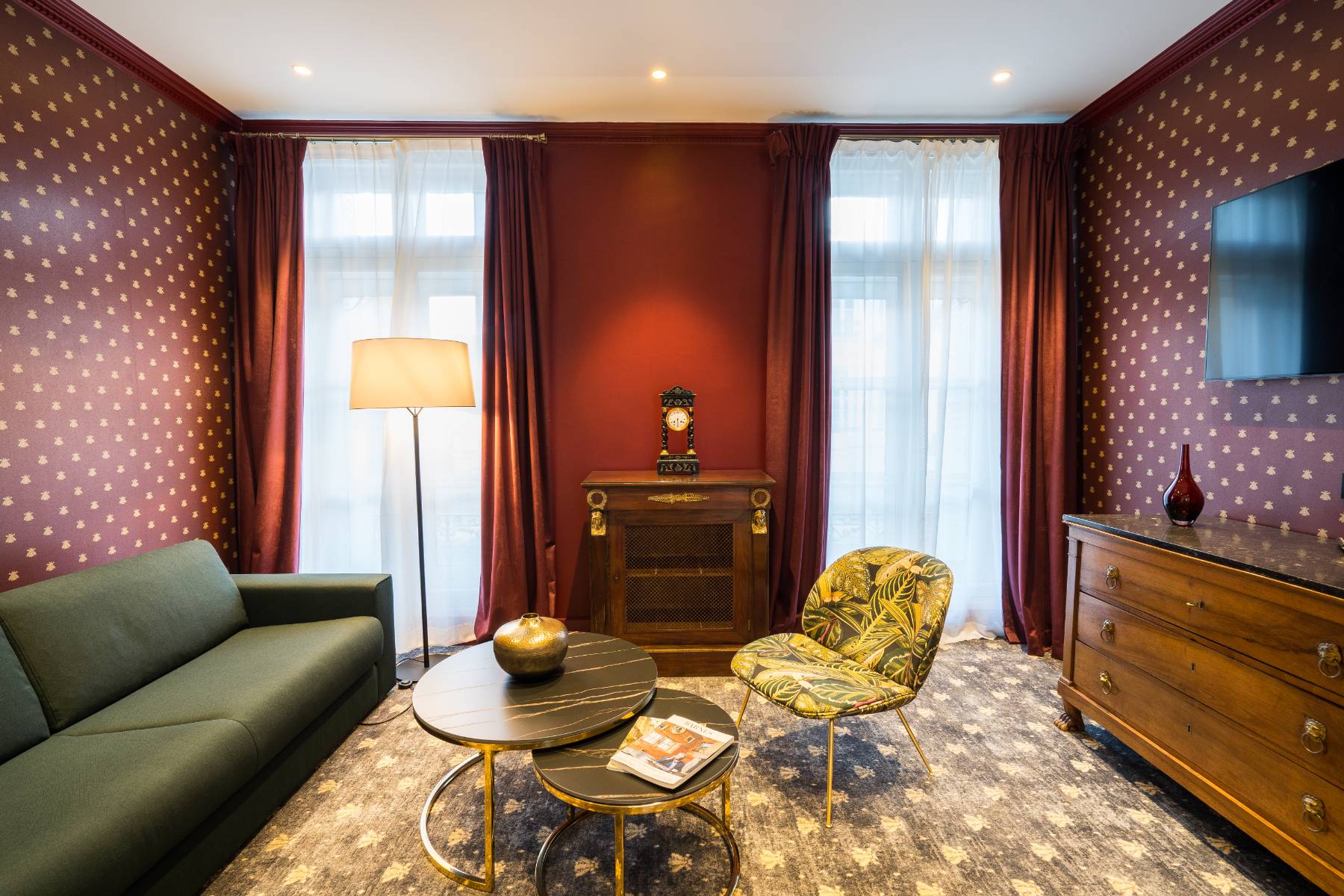 en::Living room with a large sofa, antique wooden chest with marble top, coffee tables and a plant-patterned armchair, lamp on, and large French windows with sheer curtains and drapes. fr::Salon avec grand canapé, commode ancienne en bois avec plateau en marbre, tables basses et fauteuil à motif végétal, lampe allumée, et grandes portes-fenêtres avec voilages et rideaux.