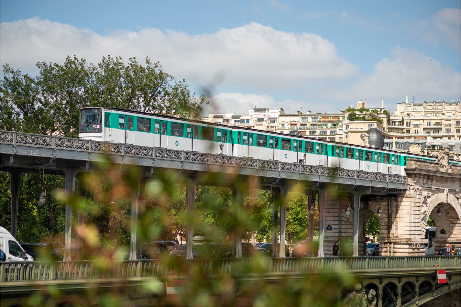 en: View of the elevated metro in Paris, with vegetation in the foreground and Haussmann-style buildings in the background. | fr: Vue du métro aérien à Paris, avec végétation au premier plan et immeubles haussmanniens en arrière-plan.