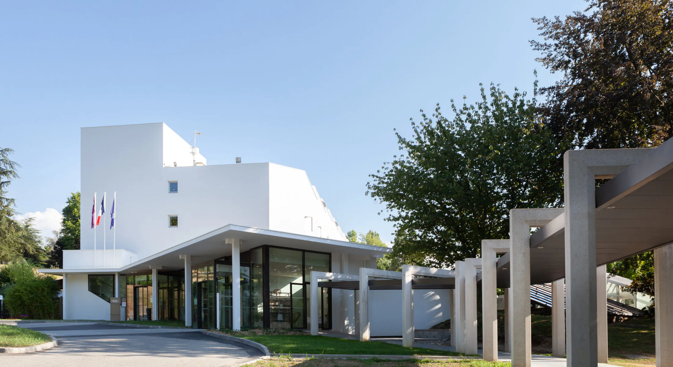 en::Modern white building with a covered wooden walkway, metal columns, large bay windows and flags. fr:: Bâtiment moderne blanc avec allée couverte en bois, colonnes métalliques, grandes baies vitrées et drapeaux.