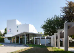 en::Modern white building with a covered wooden walkway, metal columns, large bay windows and flags. fr:: Bâtiment moderne blanc avec allée couverte en bois, colonnes métalliques, grandes baies vitrées et drapeaux.