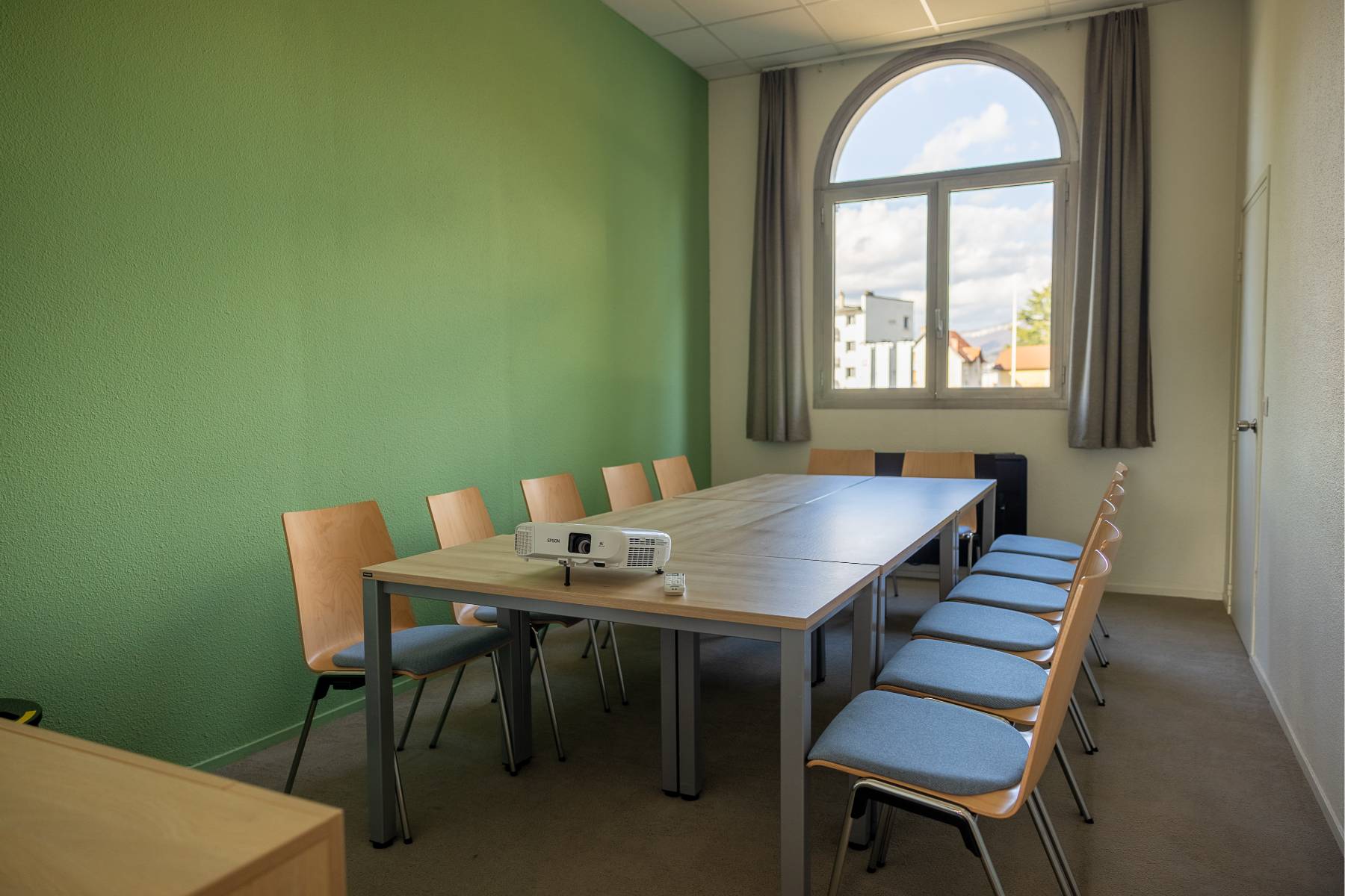 en::Rectangular wooden tables arranged in a U-shape, room with heater and air conditioning. fr::14 chaises entourant plusieurs tables en disposition board, rétro projecteur posé et orienté vers le mur