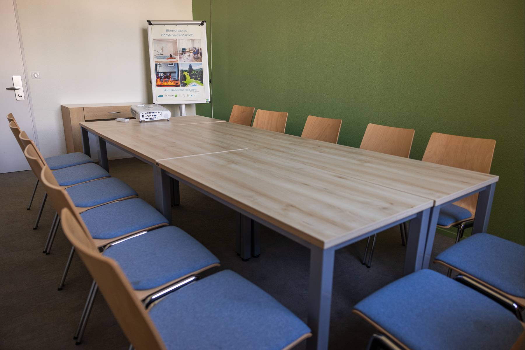 en::Rectangular wooden tables arranged in a U-shape, room with heater and air conditioning. fr::14 chaises entourant plusieurs tables en disposition board, rétro projecteur posé et orienté vers le mur