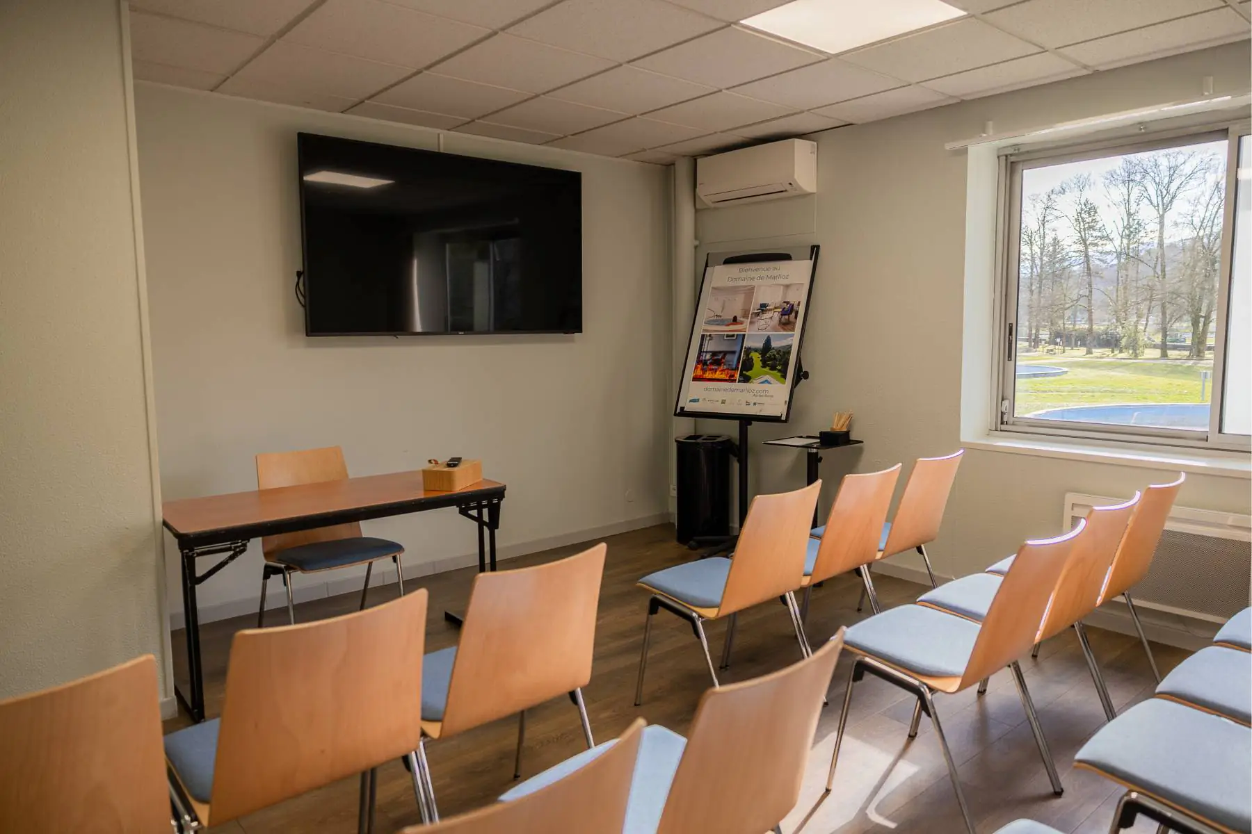 en::Desk with chair facing a row of wooden chairs with fabric seats, recycling bin, pen holder, air conditioning unit and wall-mounted TV. fr::Bureau avec chaise devant une rangée de chaises en bois avec assise tissu, poubelle de tri, pot à crayons, climatisation et écran TV mural.