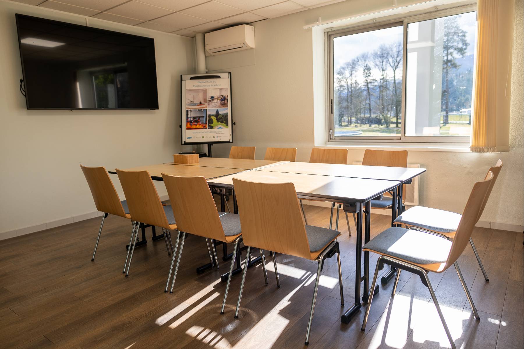 en::Four tables joined into one with wooden chairs, wall-mounted screen, window overlooking fr::Quatre tables réunies en une, chaises en bois, écran mural, fenêtre avec vue sur le parc.