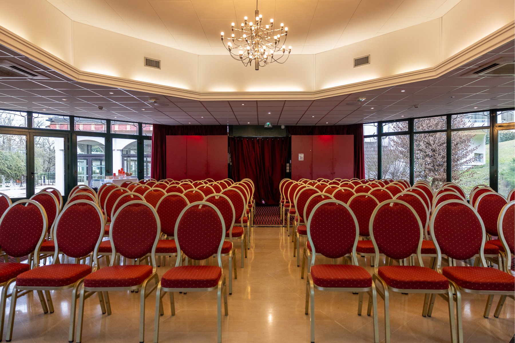 en::Rounded room with bay windows overlooking the park, chandeliers, red chairs on both sides of an aisle leading to red doors. fr::Salle arrondie entourée de baies vitrées sur le parc, lustres au plafond, chaises rouges de part et d’autre d’une allée menant à des portes rouges.