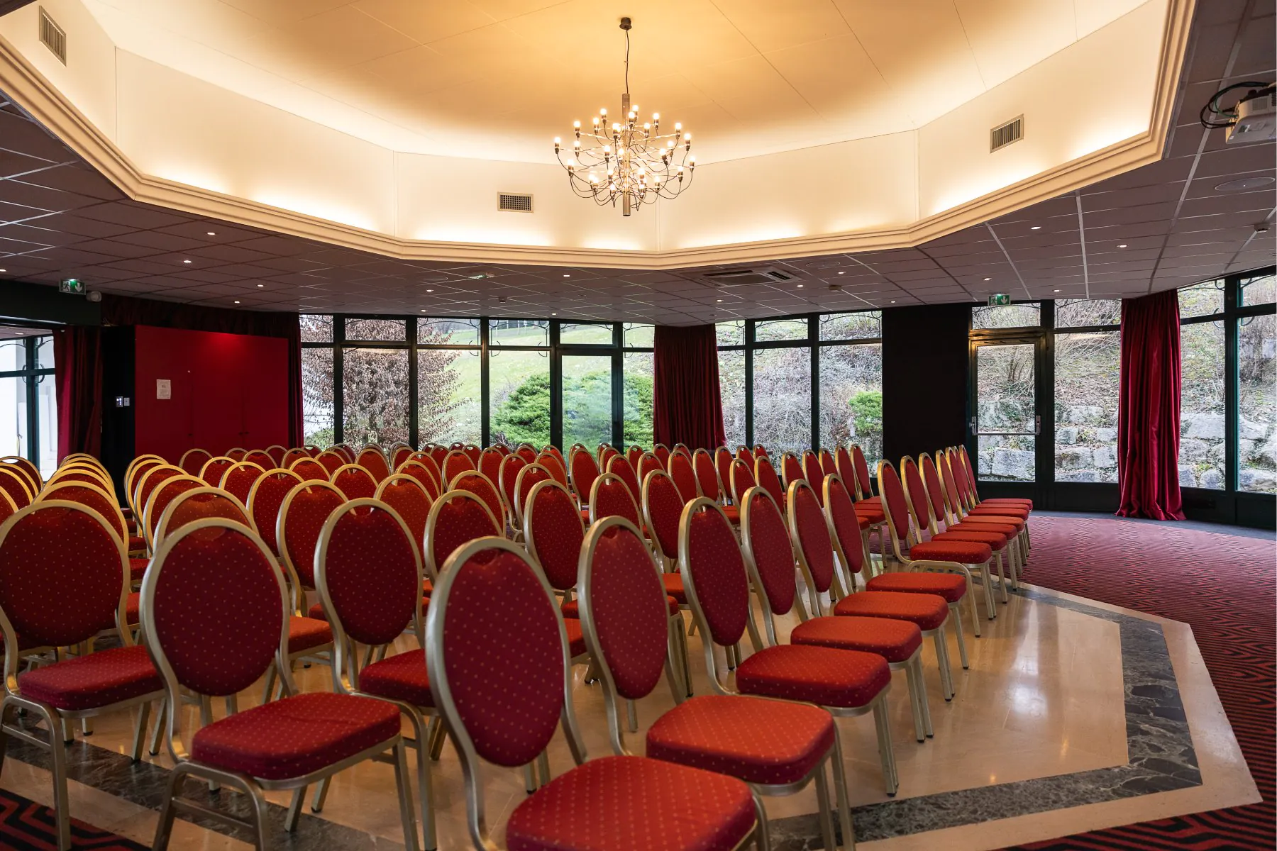 en::Meeting room under a glass roof, red theatre-style chairs, chandelier on. fr::Salle de réunion sous verrière, chaises rouges en théâtre, lustre allumé.