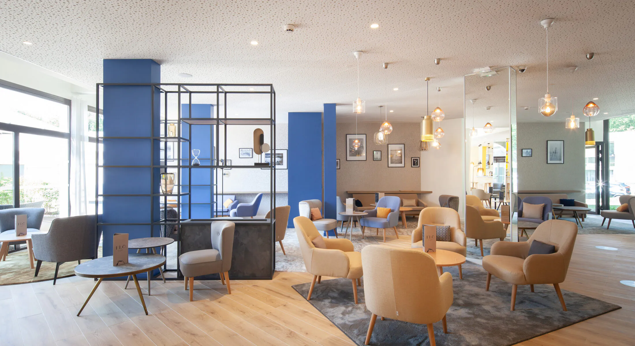 en::Lobby area with grey, blue and beige armchairs, coffee tables with menus, mirrored and pastel blue columns. fr::Espace lobby avec fauteuils gris, bleus et beiges, tables basses avec menus, colonnes miroir et bleu pastel.