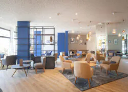 en::Lobby area with grey, blue and beige armchairs, coffee tables with menus, mirrored and pastel blue columns. fr::Espace lobby avec fauteuils gris, bleus et beiges, tables basses avec menus, colonnes miroir et bleu pastel.
