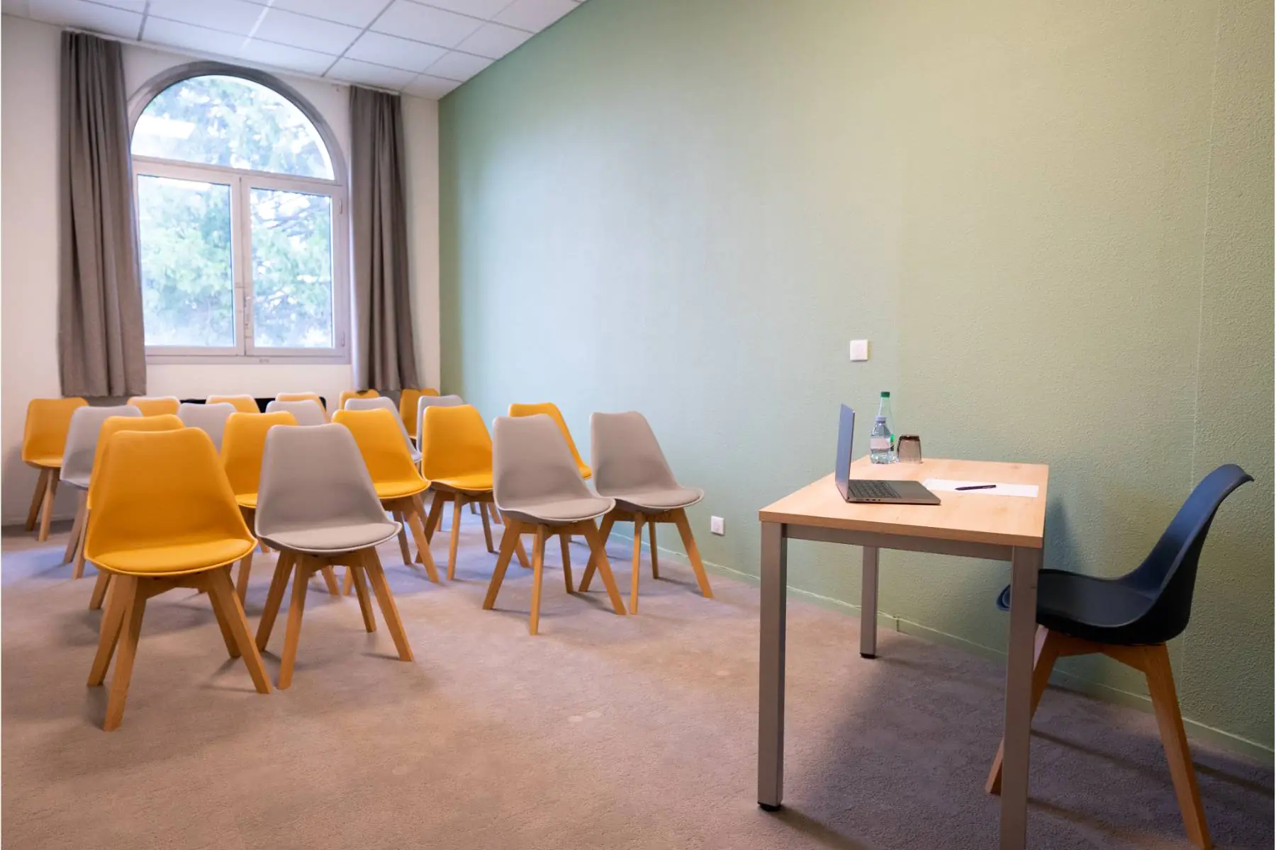 en::Desk facing rows of beige and orange chairs, window with a view of trees, beige carpet. fr::Bureau devant des rangées de chaises beiges et orangées, fenêtre avec vue sur les arbres, moquette beige.
