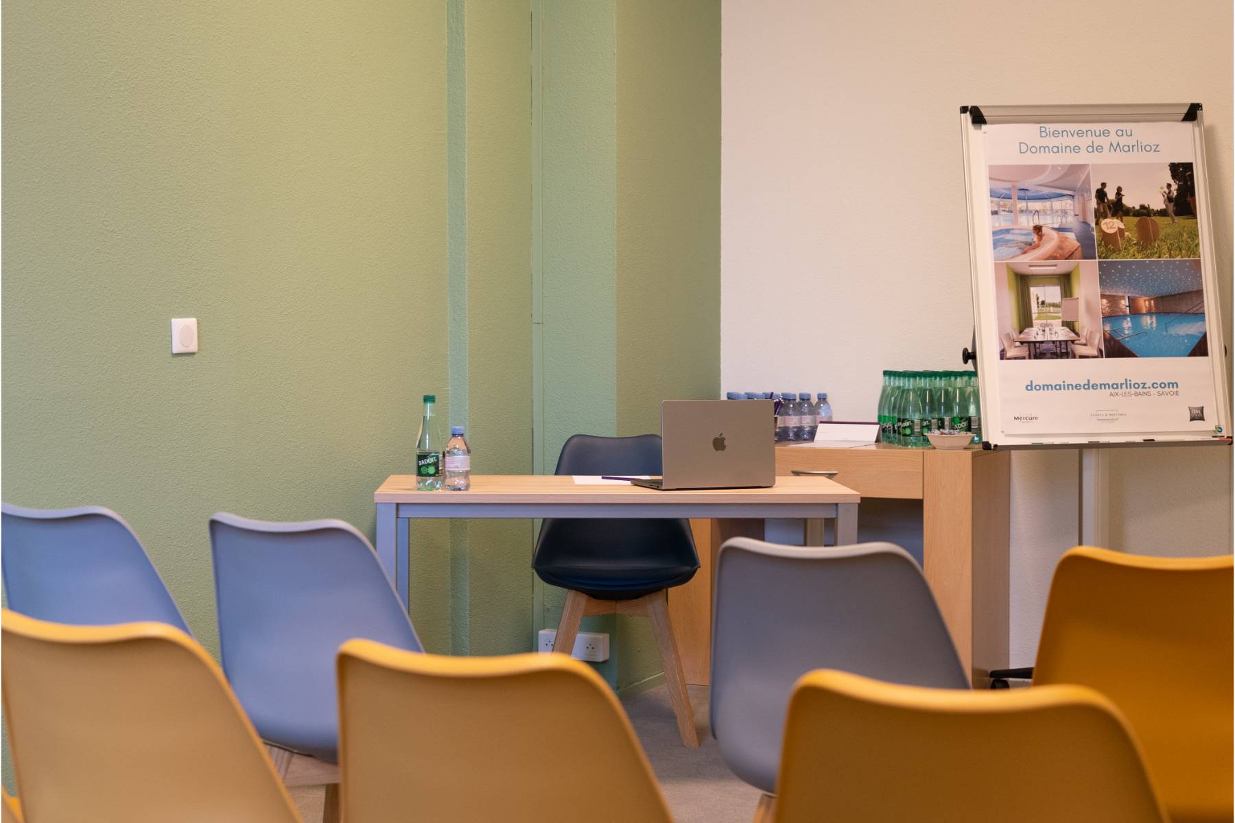 en::Desk facing rows of beige and orange chairs, cabinet with water bottles, photo taken before reduction of single-use plastics. fr::Bureau devant des rangées de chaises beiges et orangées, meuble avec bouteilles d’eau, photo prise avant réduction des plastiques.