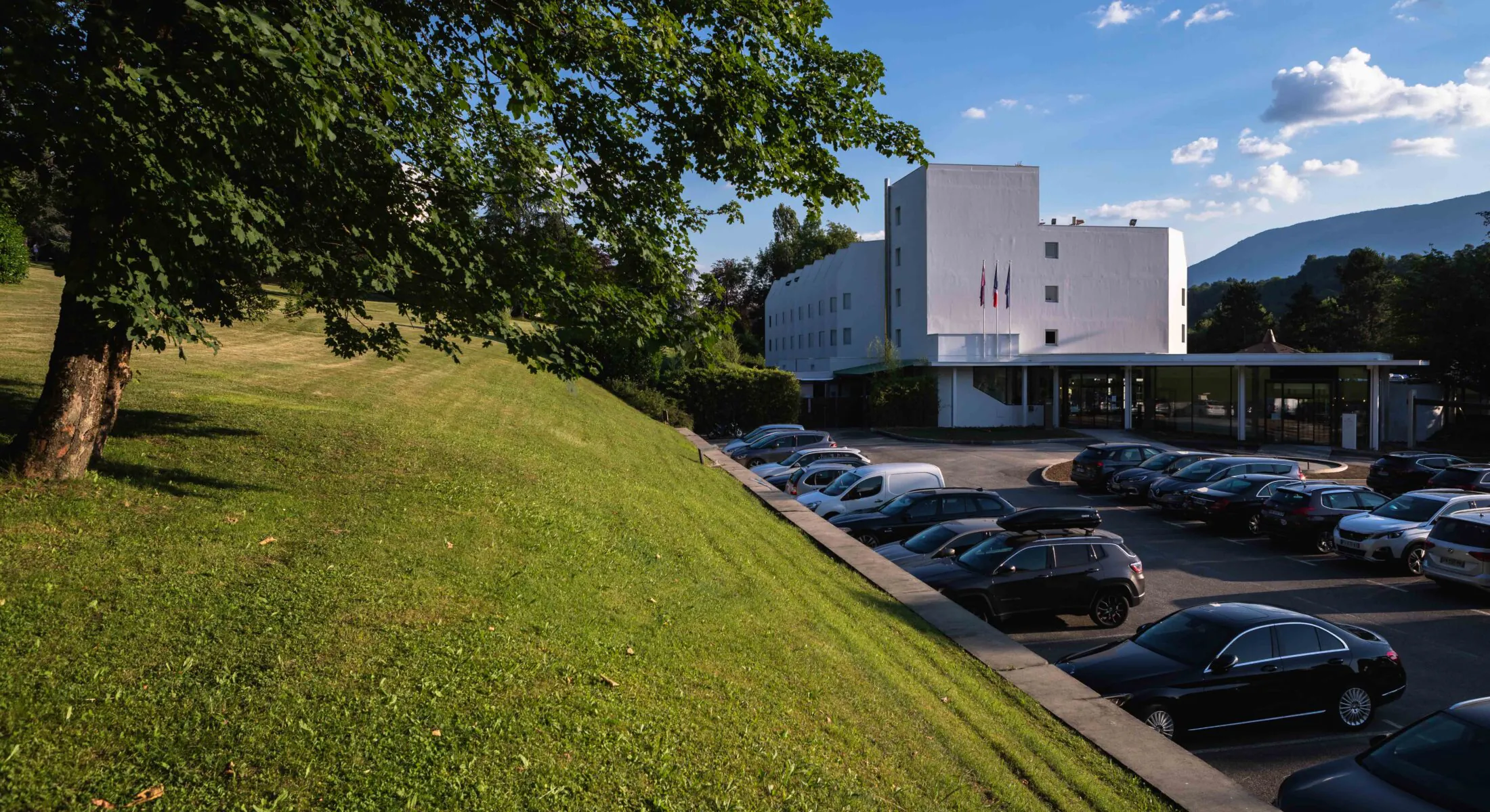 en::Modern white building with alpine mountains in the background, park in the foreground and parking area in between. fr::Bâtiment moderne blanc avec monts alpins en arrière-plan, parc au premier plan et parking entre les deux.