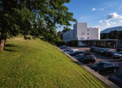 en::Modern white building with alpine mountains in the background, park in the foreground and parking area in between. fr::Bâtiment moderne blanc avec monts alpins en arrière-plan, parc au premier plan et parking entre les deux.