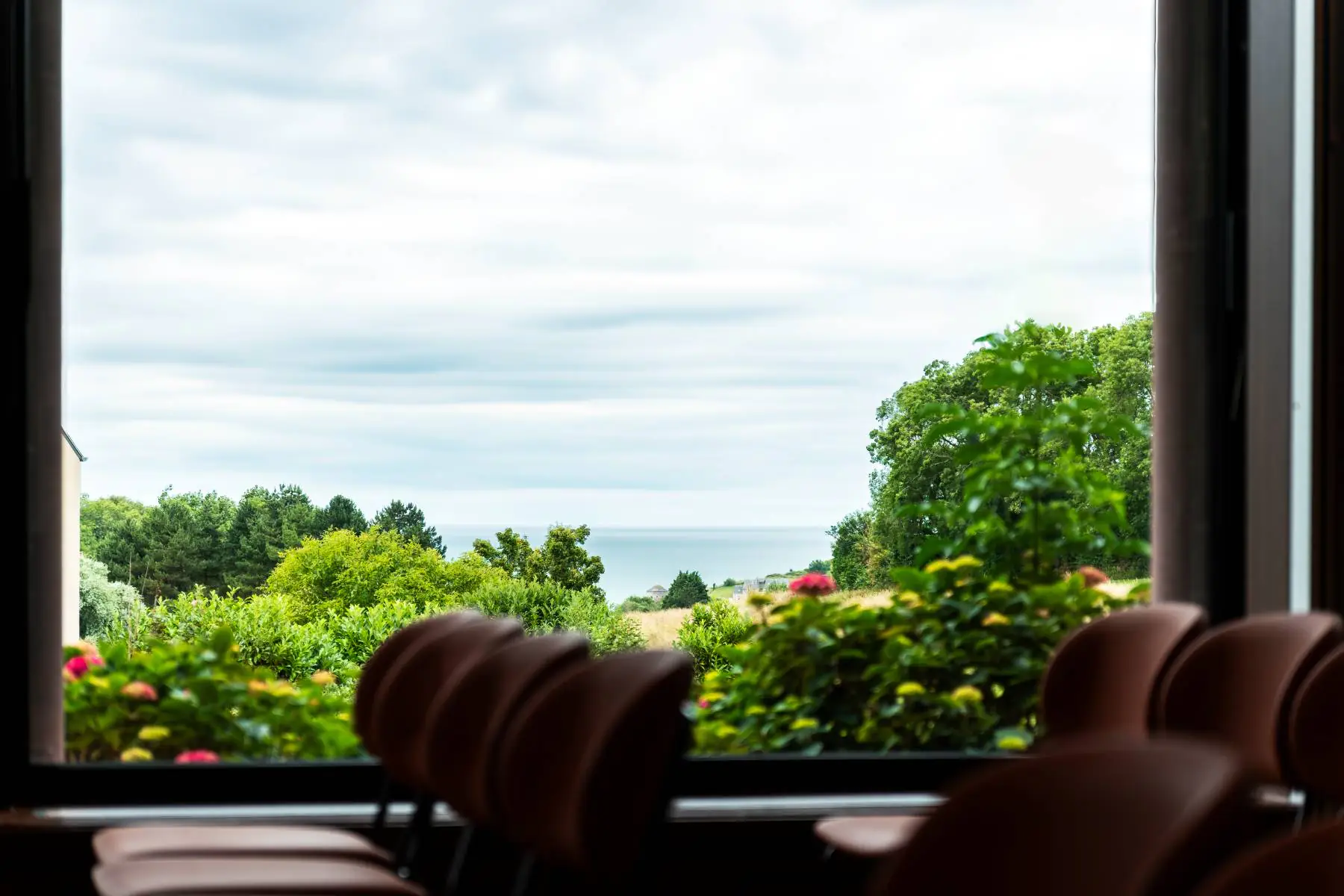 en::Row of chairs along a large window with a view of greenery, the sea, and the blue sky. fr::Rangée de chaises le long d'une baie vitrée avec vue sur la végétation, la mer, le ciel bleu.