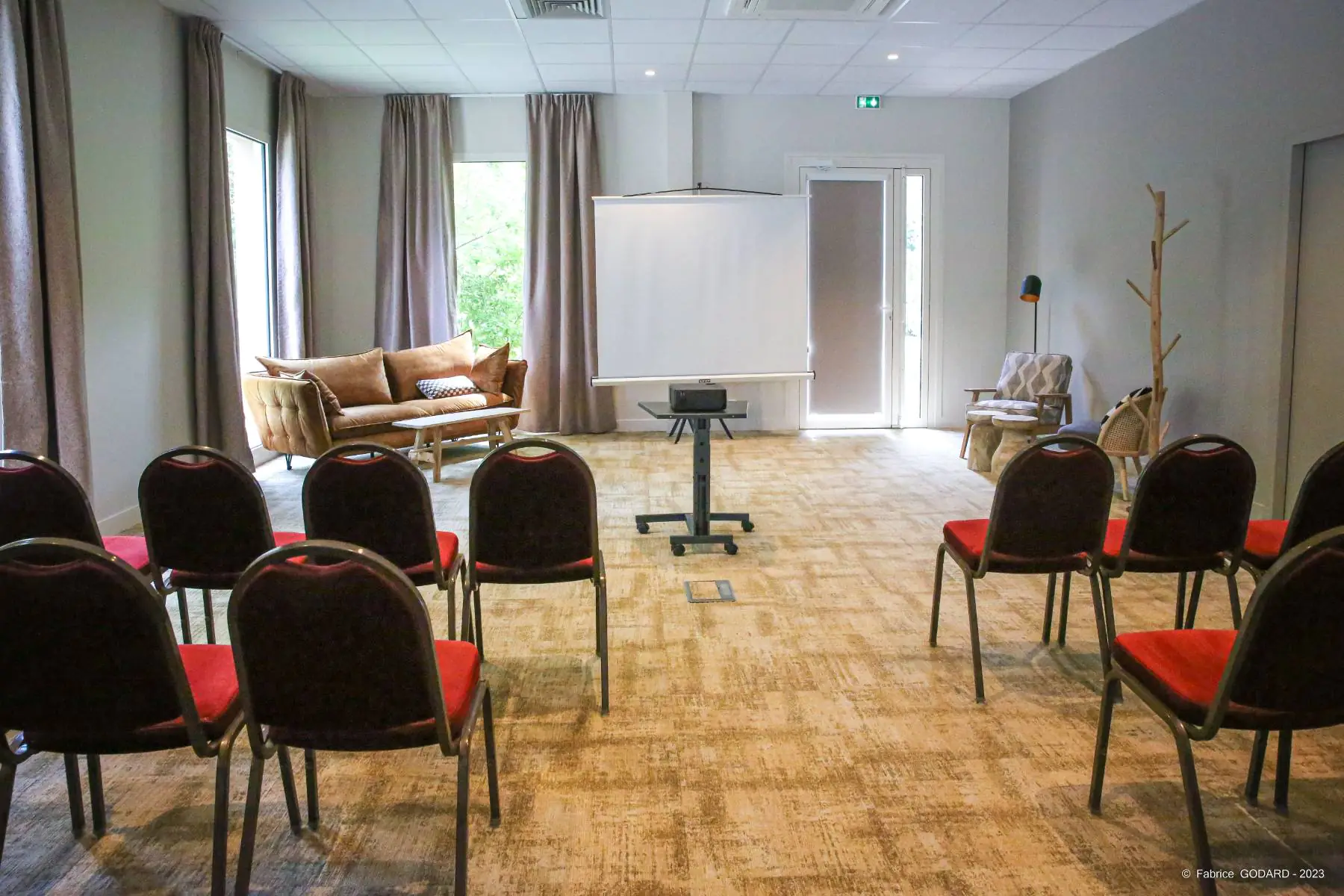 en::Beige carpet, white walls, rows of red chairs with brown frames separated by a central aisle, lounge areas with sofas around a central screen. fr::Moquette beige, murs blancs, rangées de chaises rouges avec structure marron séparées par une allée centrale, espace lounge avec canapés autour d’un écran central.