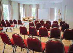 en::Rows of red chairs facing a projection screen, framed by soft pastel curtains, with a light wooden floor and sofa arranged behind the screen. fr::Rangées de chaises rouges face à un écran de projection, encadrées de doubles rideaux aux tons pastel, sur un sol en bois clair, avec canapé disposé à l’arrière de l’écran.