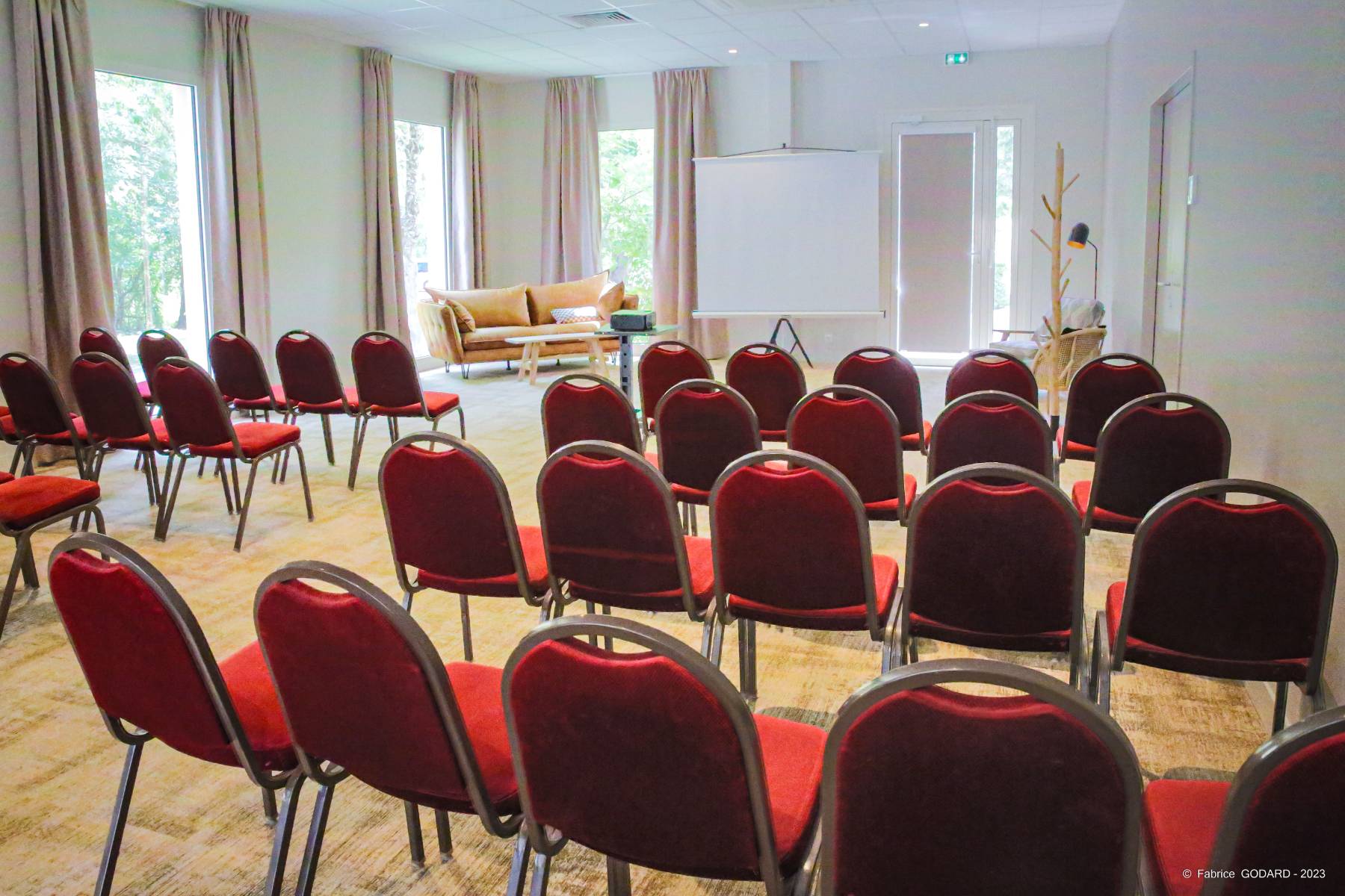 en::Rows of red chairs facing a projection screen, framed by soft pastel curtains, with a light wooden floor and sofa arranged behind the screen. fr::Rangées de chaises rouges face à un écran de projection, encadrées de doubles rideaux aux tons pastel, sur un sol en bois clair, avec canapé disposé à l’arrière de l’écran.