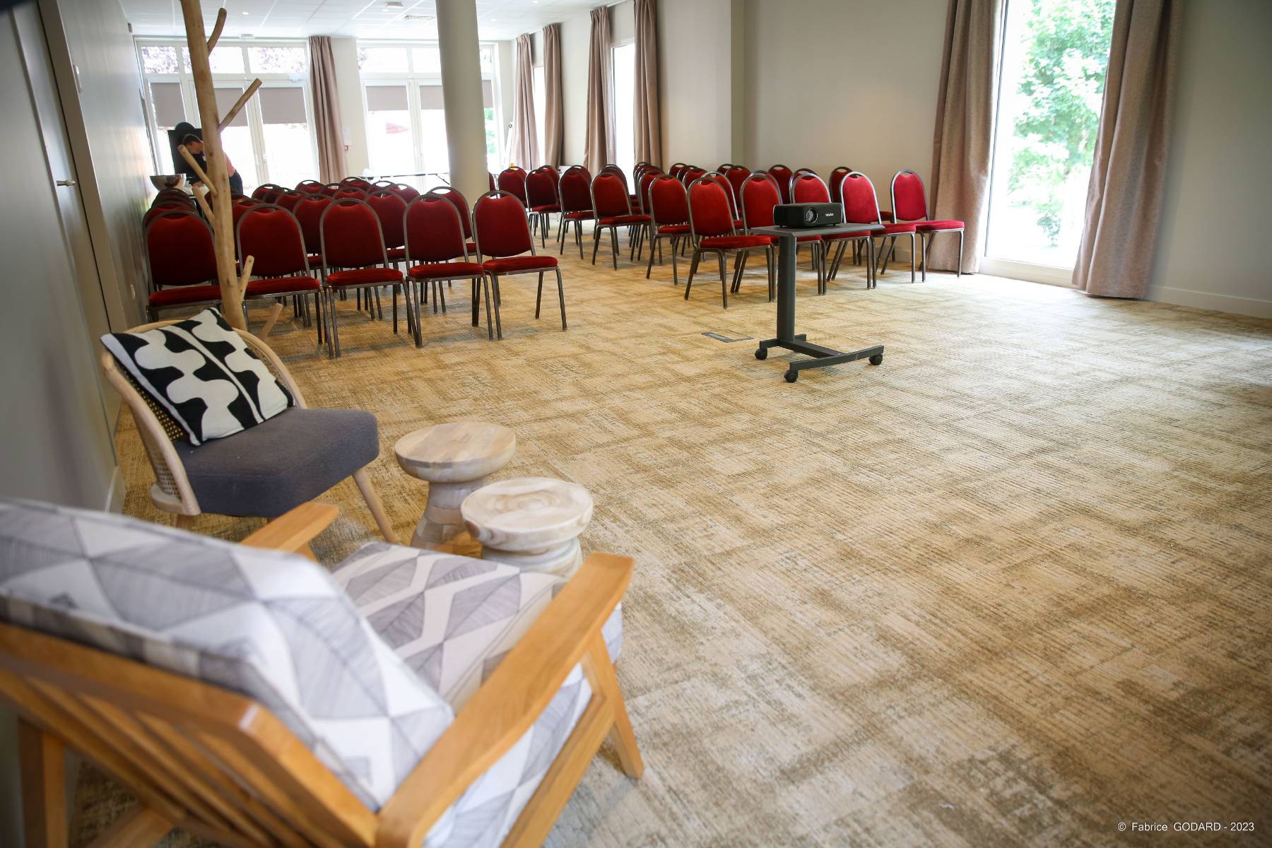 en::Beige carpet, white walls, rows of red chairs with brown frames separated by a central aisle, lounge area with sofas in one corner, large French windows and floor-to-ceiling windows. fr::Moquette beige, murs blancs, rangées de chaises rouges avec structure marron séparées par une allée centrale, espace lounge avec canapés dans un angle, grandes portes-fenêtres et baies vitrées.