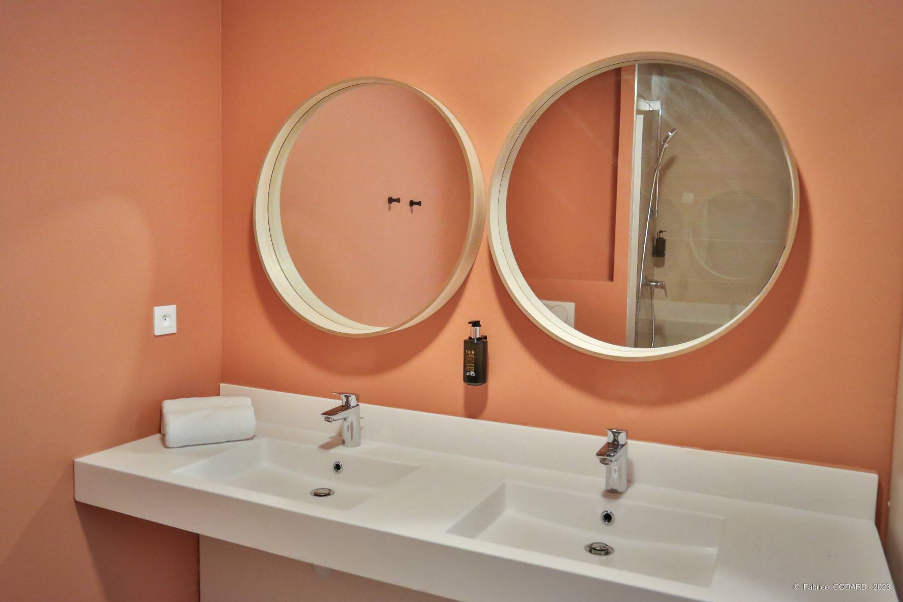 en::Bathroom with salmon-orange walls, oval mirrors and large rectangular white sink. fr::Salle de bain aux murs saumon orangé, miroirs ovales et grand lavabo blanc rectangulaire.