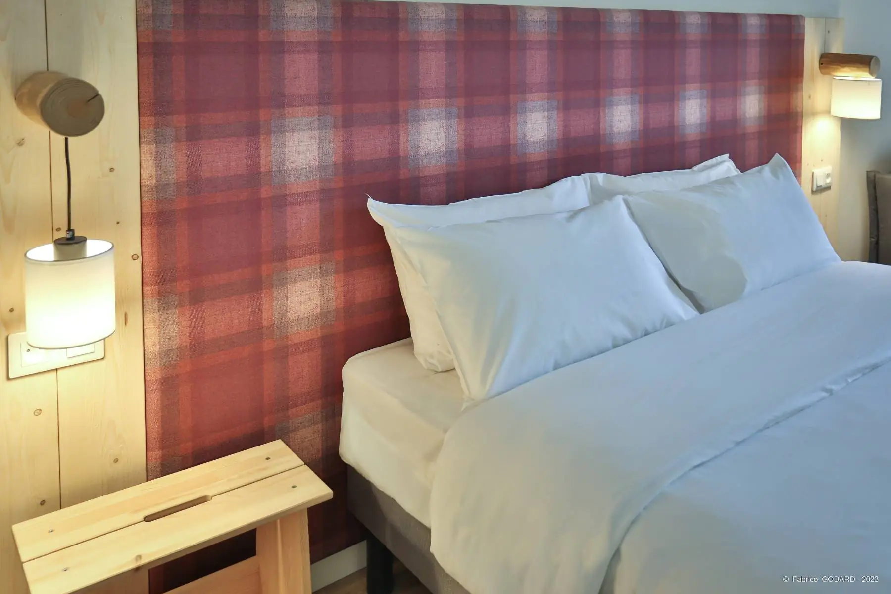 en::Double bed with wooden and checkered fabric headboard, wooden bedside tables and lights mounted on wooden logs. fr::Lit double avec tête de lit en bois et tissu à carreaux, chevets en bois et luminaires fixés sur des rondins de bois.
