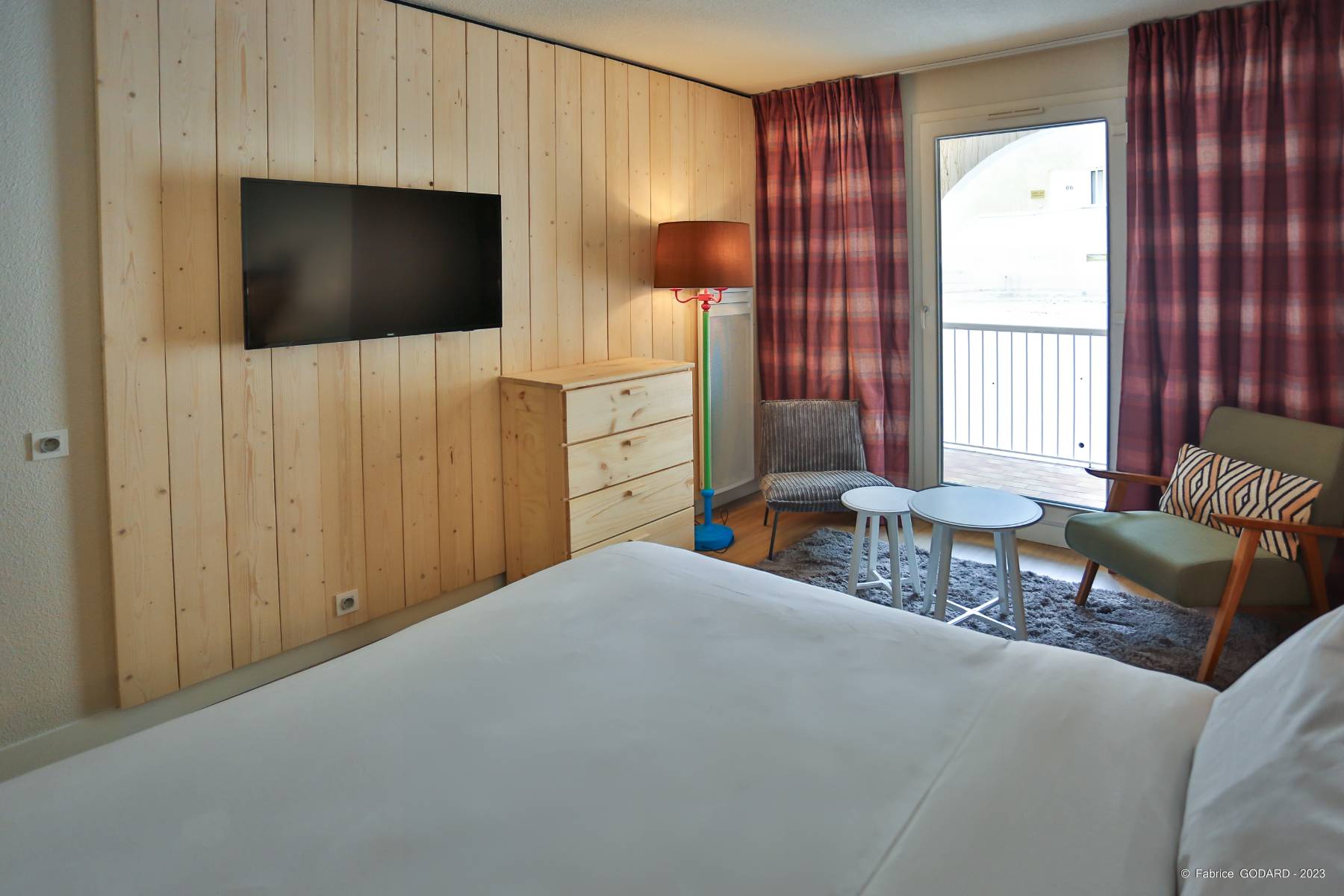 en::Double bed facing a wood-paneled wall with TV, dresser and radiator, lounge area near the balcony. fr::Lit double face à un mur habillé de bois avec TV, commode et radiateur, coin lounge près du balcon.