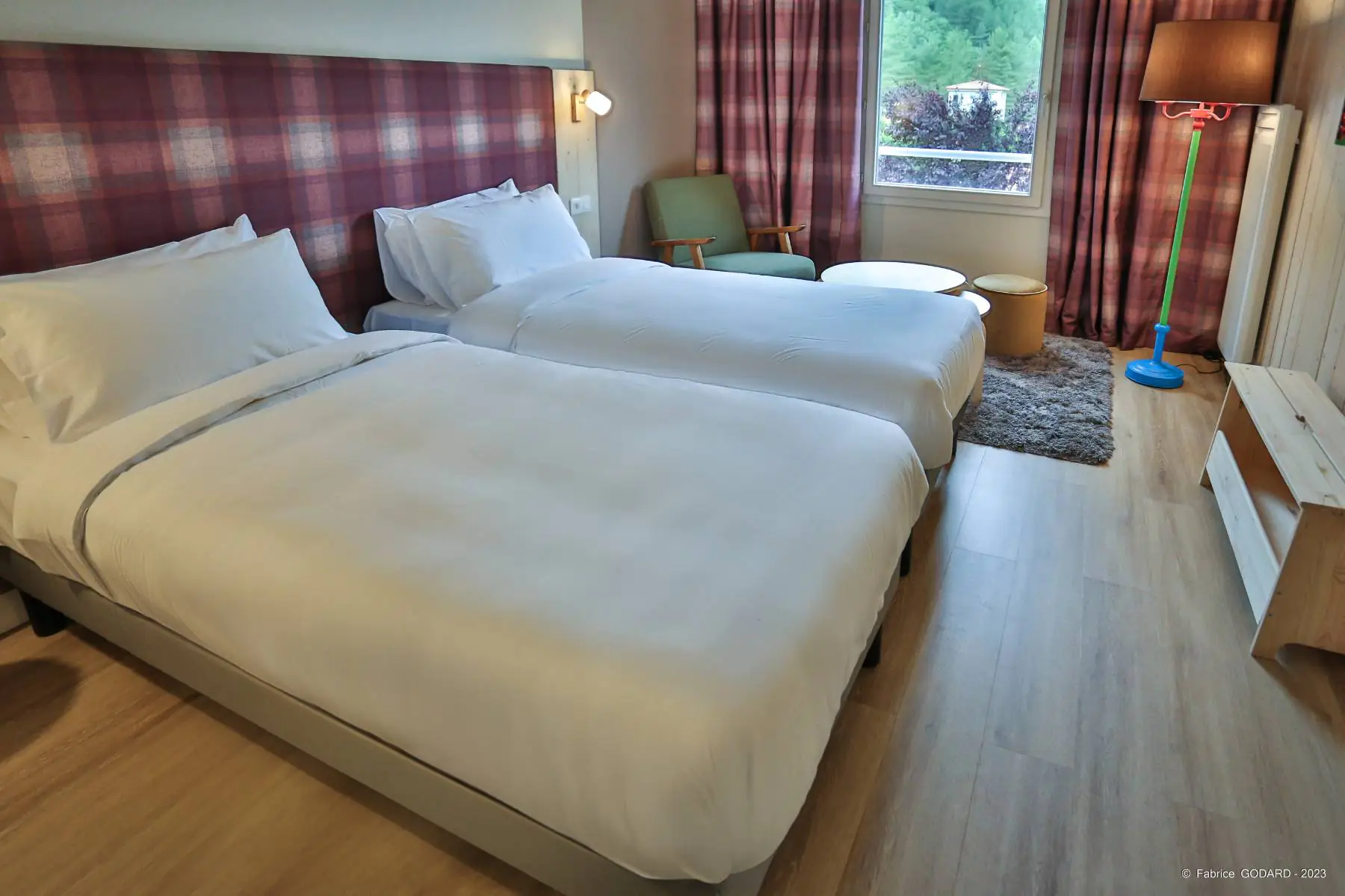 en::Twin beds with checkered fabric headboards, green and blue lamp with brown lampshade, wooden luggage rack, grey rug and nature view. fr::Lits jumeaux avec têtes de lit en tissu à carreaux, lampe au pied vert et bleu avec abat-jour marron, porte-valise en bois, tapis gris et vue sur la nature.