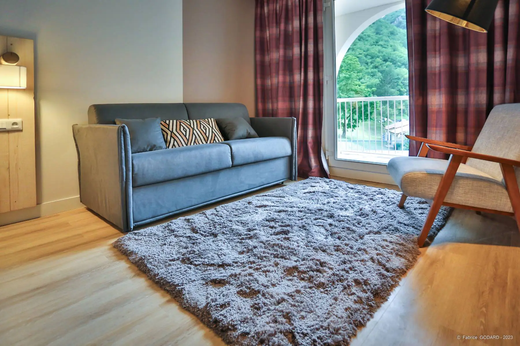 en::Grey double sofa bed with cushions, grey rug, upholstered chair with wooden armrests and legs, with a view of nature from the accessible balcony. fr::Canapé-lit double gris avec coussins, tapis gris, chaise en tissu avec accoudoirs et pieds en bois, vue sur la nature depuis le balcon accessible.