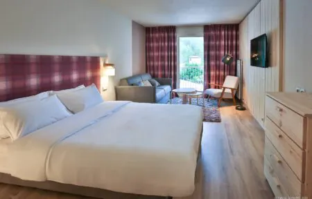 en::Large double bed with red and blue checkered headboard, matching curtains open onto mountain vegetation, wood wall, wooden dresser and grey sofa. fr::Grand lit double avec tête de lit à carreaux rouges et bleus, rideaux assortis ouverts sur la végétation montagnarde, mur en bois, commode en bois et canapé gris.