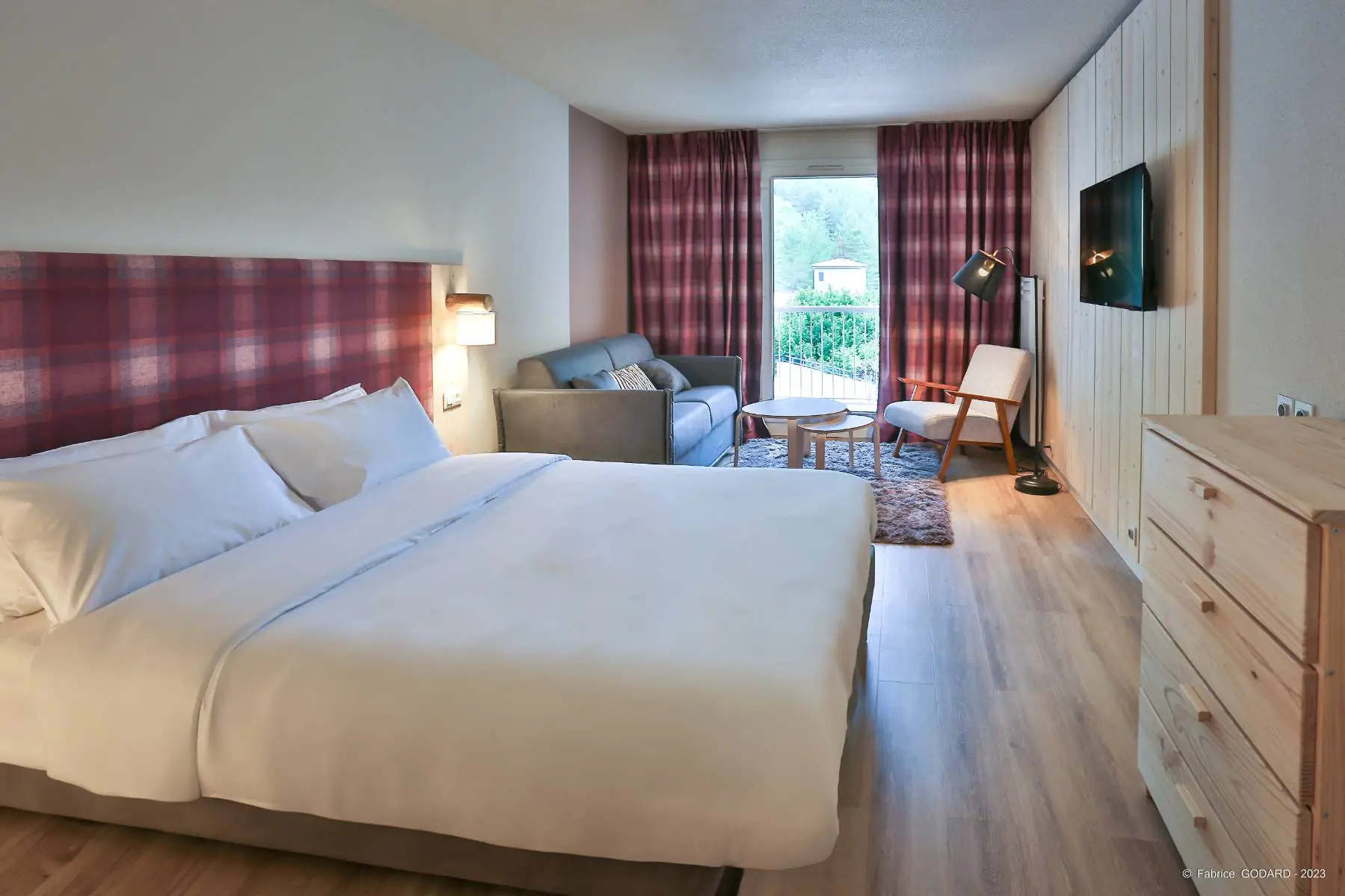 en::Large double bed with red and blue checkered headboard, matching curtains open onto mountain vegetation, wood wall, wooden dresser and grey sofa. fr::Grand lit double avec tête de lit à carreaux rouges et bleus, rideaux assortis ouverts sur la végétation montagnarde, mur en bois, commode en bois et canapé gris.