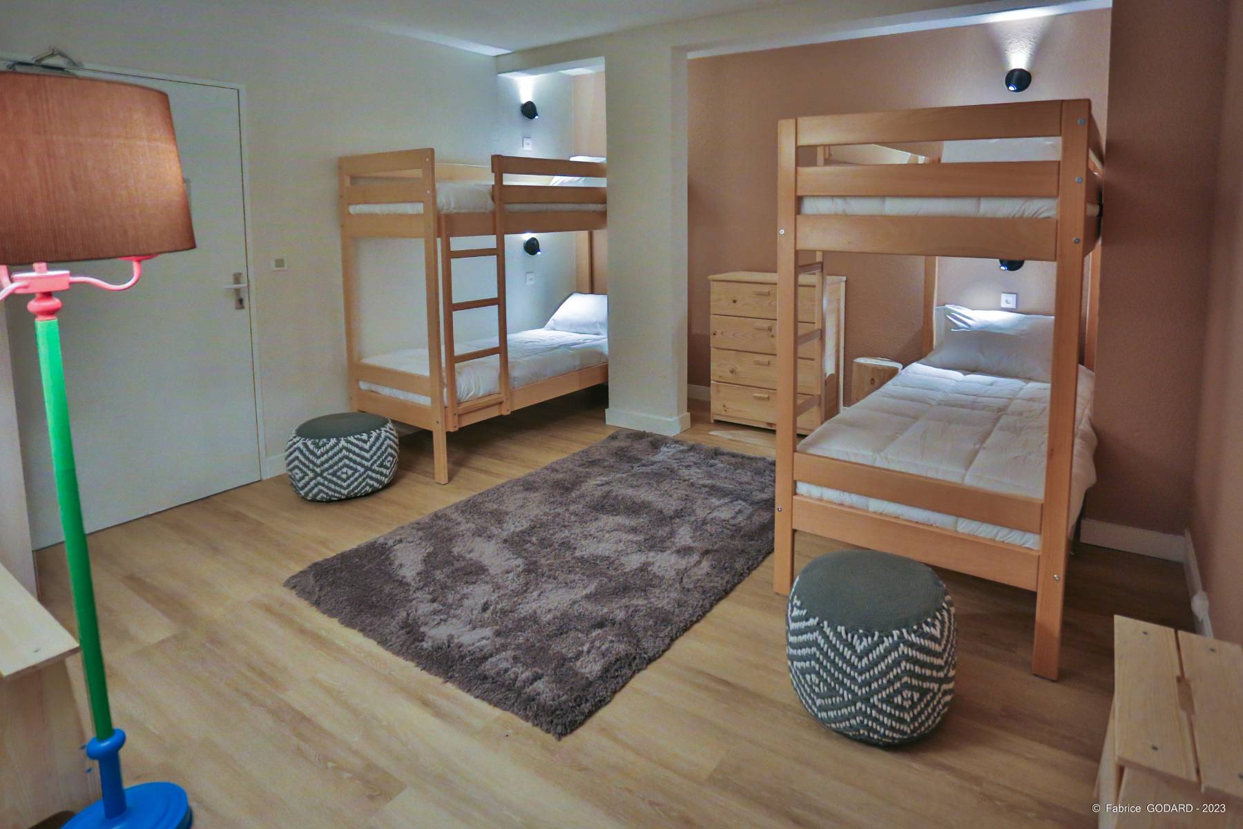 en::Two sets of bunk beds in a bedroom, rug and dresser between them, matching poufs and colorful lamp. fr::Deux ensembles de lits superposés dans une chambre, tapis et commode entre les deux, poufs assortis et lampe colorée.