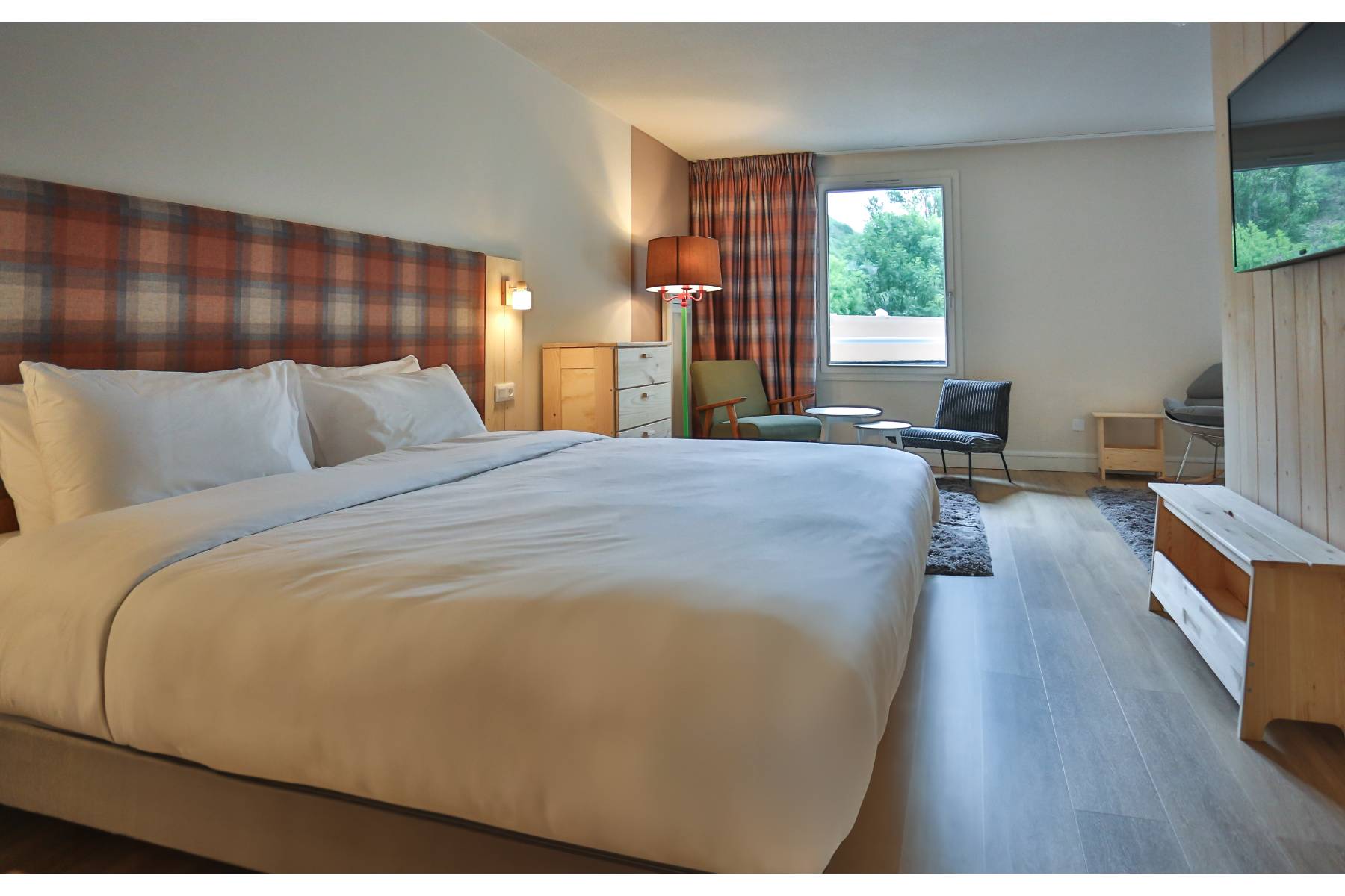 en::Double bed facing a wood-paneled wall with TV and wooden bench, lounge area near the window. fr::Lit double devant un mur en lambris avec télévision et banc en bois, coin lounge près de la fenêtre