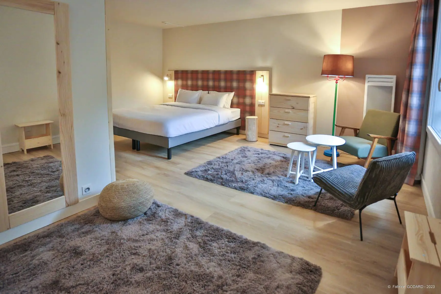 en::Bedroom with wooden wall mirror, double bed and lounge area, wooden flooring and rug. fr::Chambre avec miroir mural en bois, lit double et espace salon, parquet et tapis au sol.