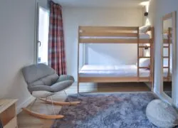 en::Wooden bunk beds facing a large window, soft grey rug, grey rocking chair with wooden legs and wooden mirror. fr::Lits superposés en bois face à une grande fenêtre, tapis gris moelleux, chaise à bascule grise avec pieds en bois et miroir en bois.