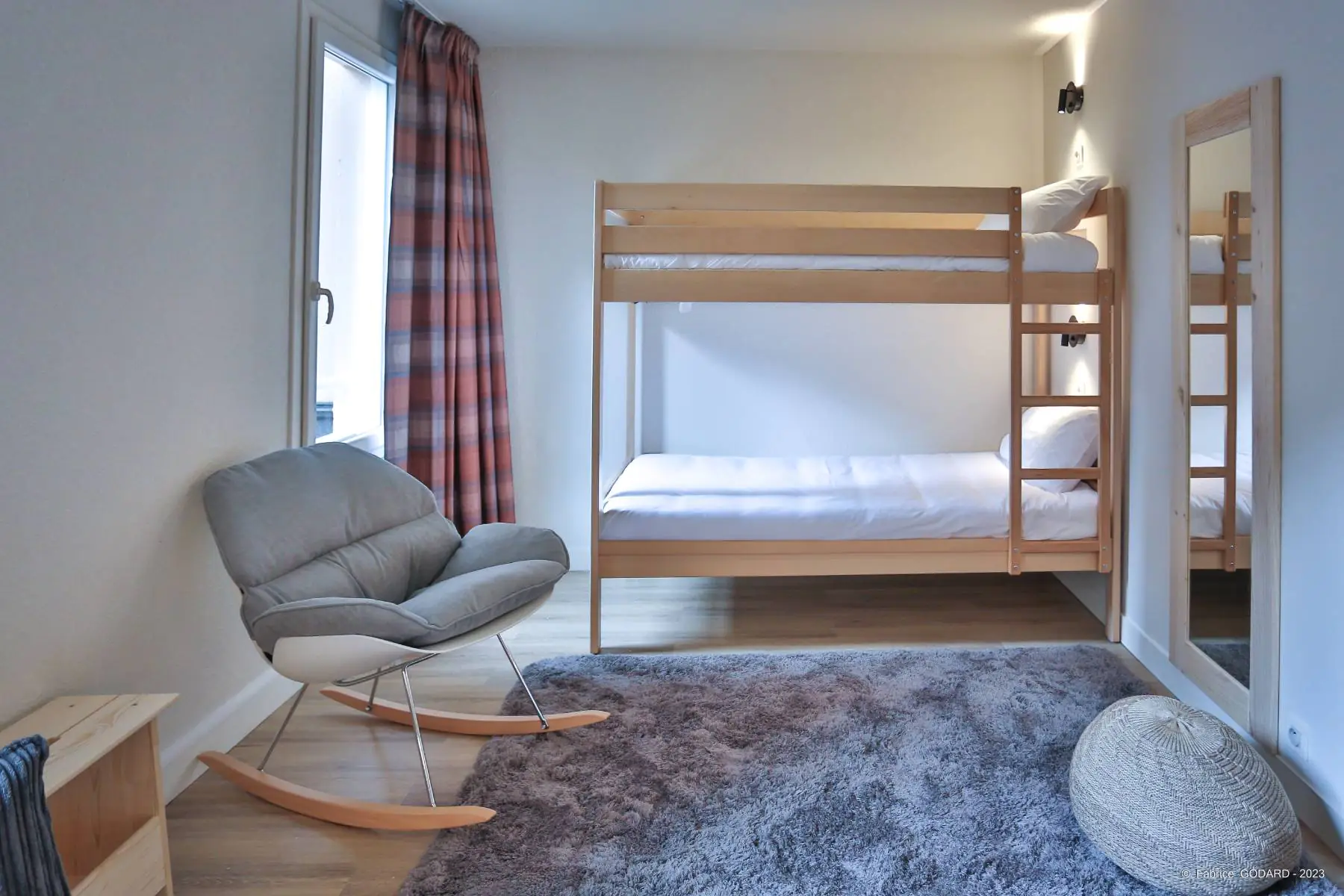 en::Wooden bunk beds facing a large window, soft grey rug, grey rocking chair with wooden legs and wooden mirror. fr::Lits superposés en bois face à une grande fenêtre, tapis gris moelleux, chaise à bascule grise avec pieds en bois et miroir en bois.
