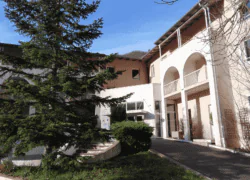 en::White and beige hotel façade with a large pine tree at the entrance, mountains visible above the rooftops. fr::Façade d’hôtel blanche et beige, sublimée par un grand sapin à l’entrée, avec les montagnes en toile de fond au-dessus des toits.