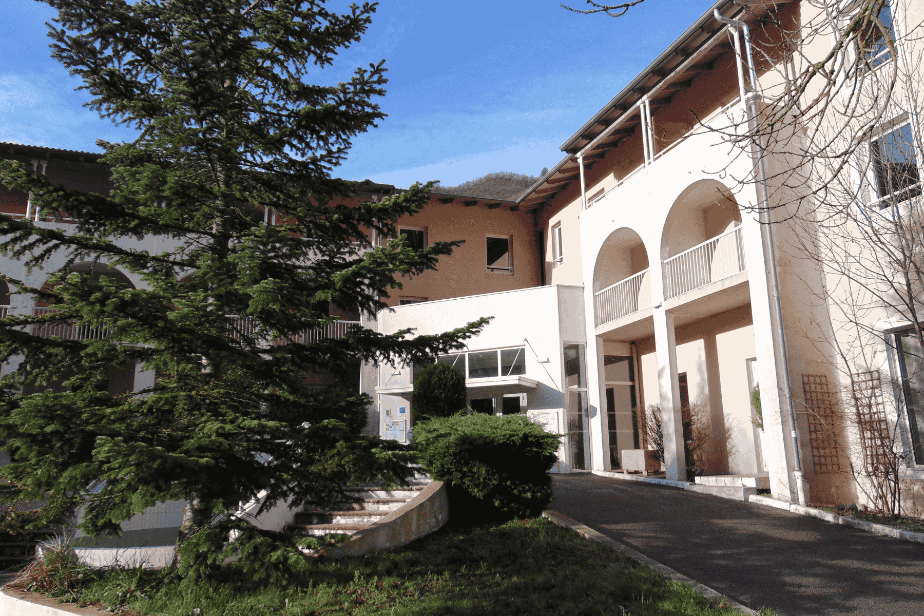 en::White and beige hotel façade with a large pine tree at the entrance, mountains visible above the rooftops. fr::Façade d’hôtel blanche et beige, sublimée par un grand sapin à l’entrée, avec les montagnes en toile de fond au-dessus des toits.