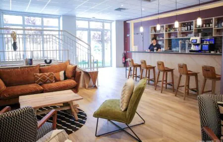 en::Wood-clad bar near large windows overlooking trees and lawn, facing a white staircase, lounge area with rustic coffee tables and mixed seating in various colors and shapes. fr::Bar en bois près des baies vitrées avec vue sur les arbres et la pelouse, face à un escalier blanc, espace lounge avec tables basses rustiques et assises variées.