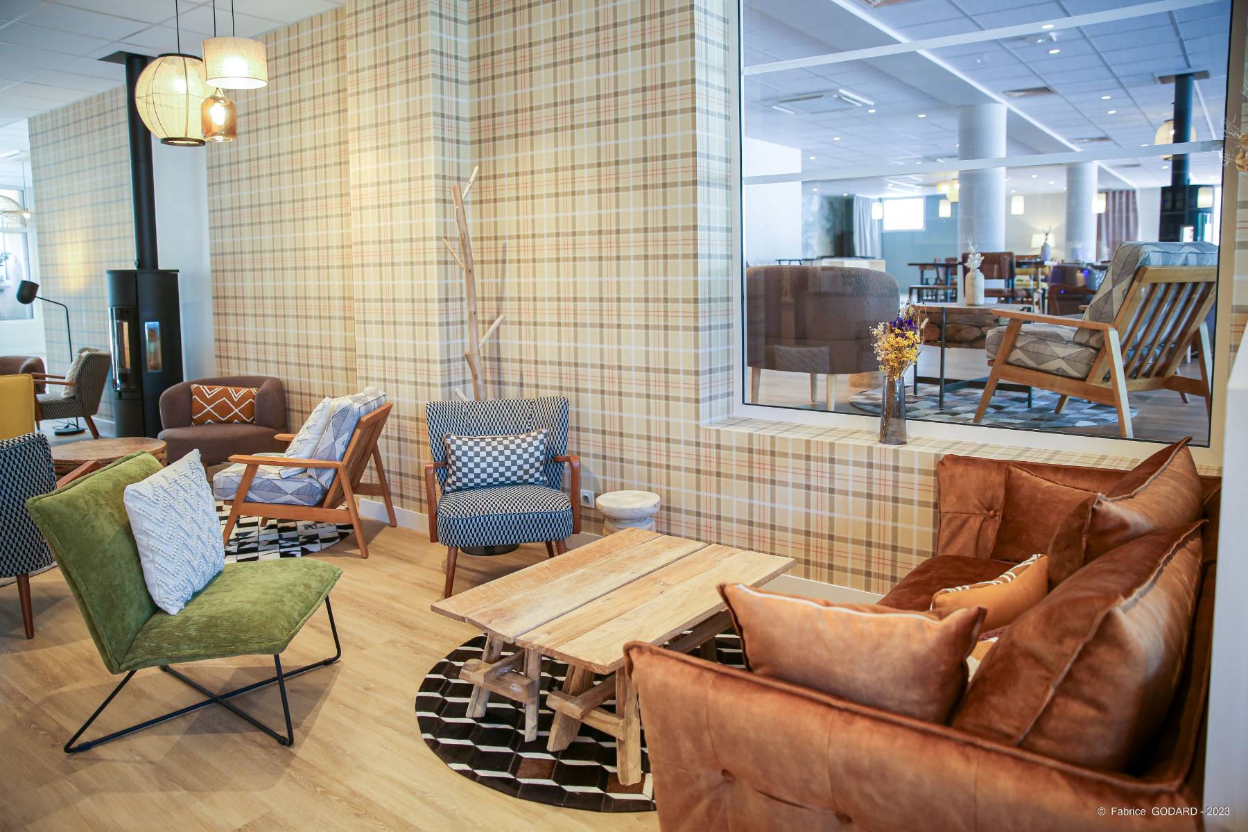 en::Wood-clad bar near large windows overlooking trees and lawn, facing a white staircase, lounge area with rustic coffee tables and mixed seating, tree trunk used as a coat rack, stove heater and wall opening with a glimpse into the restaurant. fr::Espace lounge avec tables basses rustiques et assises variées, tronc d’arbre faisant office de porte-manteau, poêle de chauffage et ouverture murale avec aperçu sur la salle de restaurant.