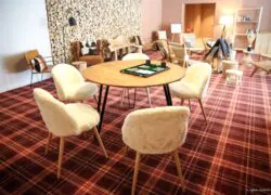 en::Burgundy and orange checkered carpet, round wooden table with board games, surrounded by soft white upholstered chairs, lounge area in the background with a bookcase and mountain-style décor. fr::Moquette à carreaux bordeaux et orange, table ronde en bois avec jeux de société, entourée de chaises au revêtement blanc moelleux, salon en arrière-plan avec bibliothèque et décor montagnard.